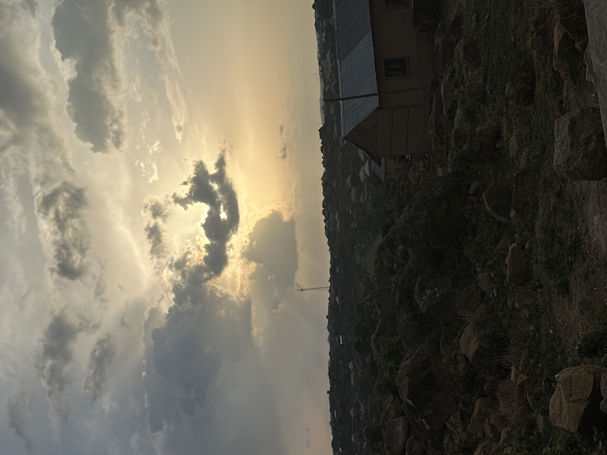 The image shows a landscape in Jos, Nigeria, under a cloudy, golden-lit sky. The right side of the image features a rocky hillside with sparse vegetation. On the upper right, there is a small building with a gabled roof. Down the hill, smaller structures and buildings are visible. A tall telecommunications tower is seen in the distance near the center of the frame. The sky takes up the left side of the image, filled with dark and light gray clouds and a golden glow near the horizon.