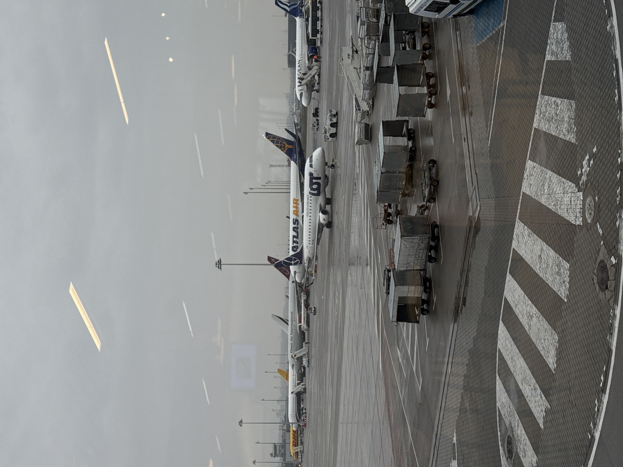 The image depicts an airport scene, specifically Sofia Airport in Bulgaria. The view is from inside a terminal, looking out onto the tarmac. Several airplanes are visible, including an LOT Polish Airlines plane. There are cargo carts lined up next to the planes. The weather appears overcast. The ground is wet, suggesting it has recently rained. The image also captures a portion of a zebra-striped walkway and a paved area. The light is somewhat diffused, possibly due to the weather conditions.