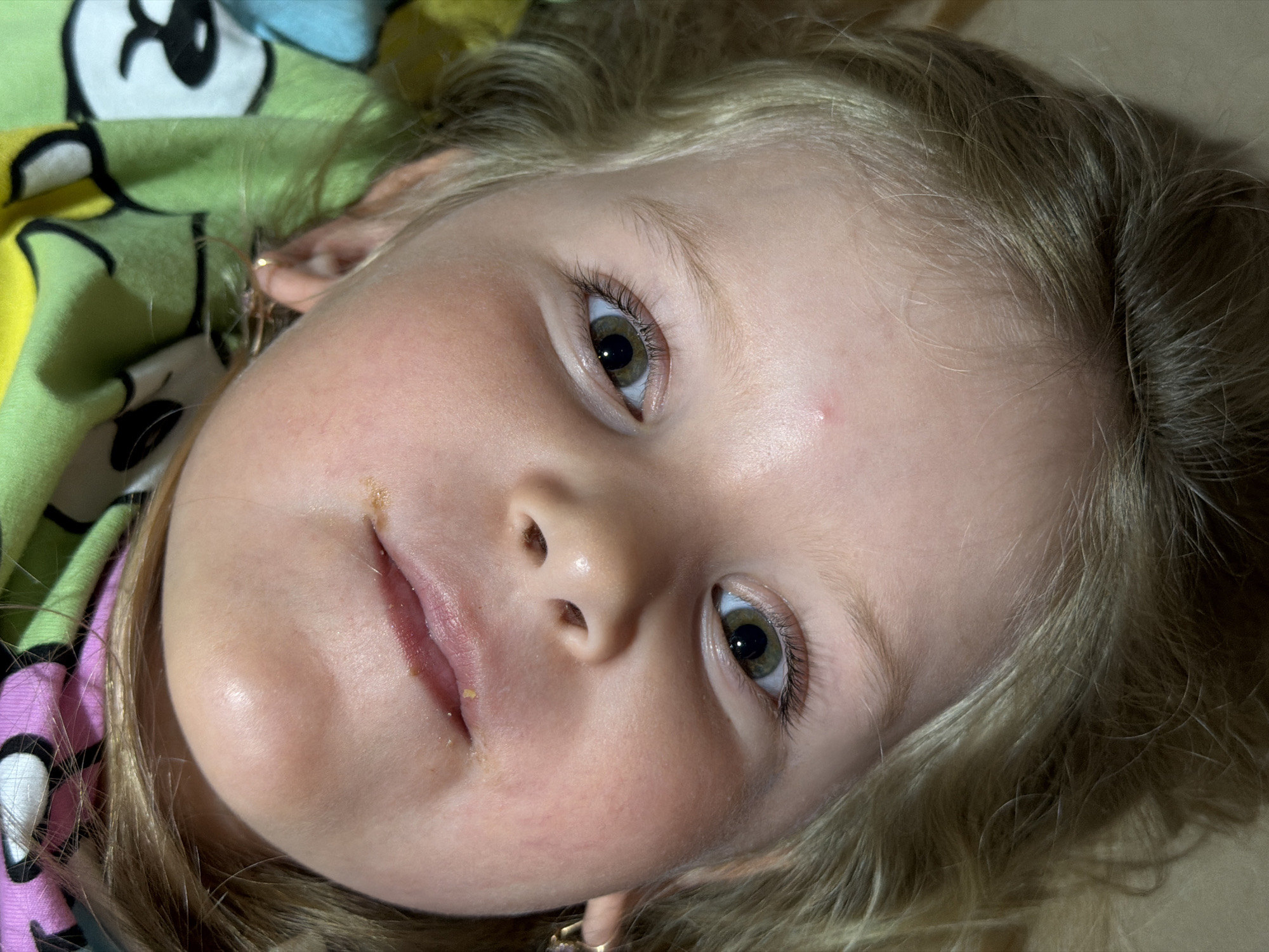 The specific location within the city of Chaikovsky, Russia, cannot be determined from the provided image.
The image presents a close-up, inverted view of a young, fair-skinned individual, likely female, with blonde hair. The subject's head is oriented upside down within the frame. Both eyes are open, with pupils moderately dilated, and the irises exhibit a hazel (green-brown) coloration, directed forward towards the viewer. The nose is small with visible nostrils. The lips are slightly parted, and small, irregular yellowish particulate matter, consistent with food residue, is present on the lower lip and surrounding skin. A minor reddish blemish is visible on the upper left cheek/forehead area (viewer's left). Portions of both ears are visible, each adorned with a small, circular, metallic earring, possibly gold-toned. The blonde hair appears fine, somewhat disheveled, and spreads around the head, with darker roots visible in some areas. The subject is situated against a soft textile background featuring a colorful, patterned design, including blocks of light green, pink, and yellow, with black-outlined cartoon-like figures. This textile suggests a garment, blanket, or bedding. The lighting is soft and even, indicating an indoor or shaded environment.