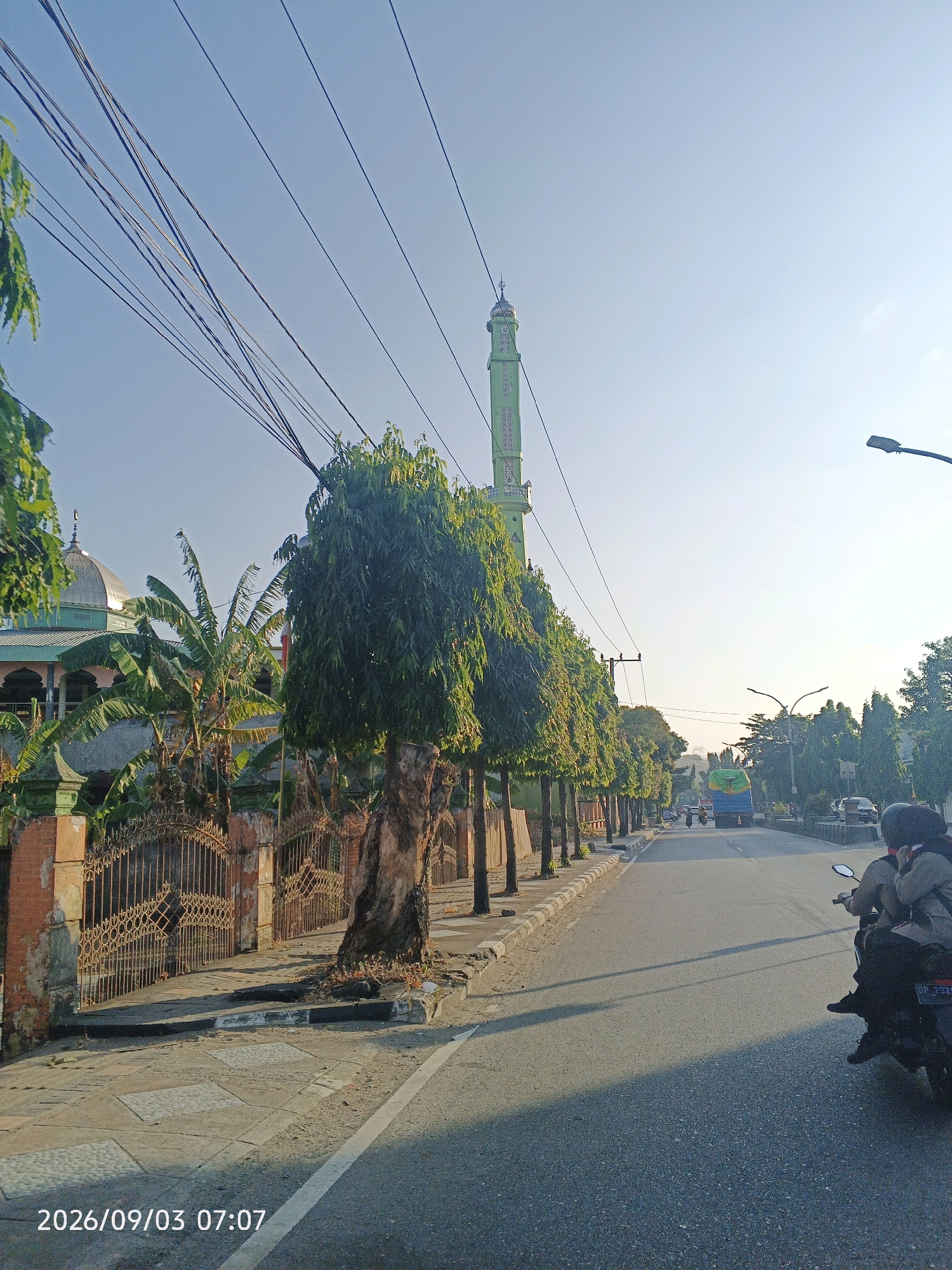 The image depicts a street scene in Pare-Pare, Indonesia, likely during the early morning based on the timestamp and lighting. On the left side of the street, a mosque with a dome and a tall, slender minaret is visible, surrounded by lush green trees, including banana plants and other foliage. A decorative metal fence borders the mosque property. A paved sidewalk runs alongside the street, with trees lining its edge, casting long shadows.
The street itself is asphalt, with a white line marking the lane. Several vehicles are present, including a blue truck in the distance, other cars, and a motorcycle in the foreground. Two individuals are riding on the motorcycle, one driving and the other as a passenger, both wearing helmets. Utility lines crisscross the sky above the scene. The overall environment appears to be a well-trafficked urban or suburban area with significant greenery.