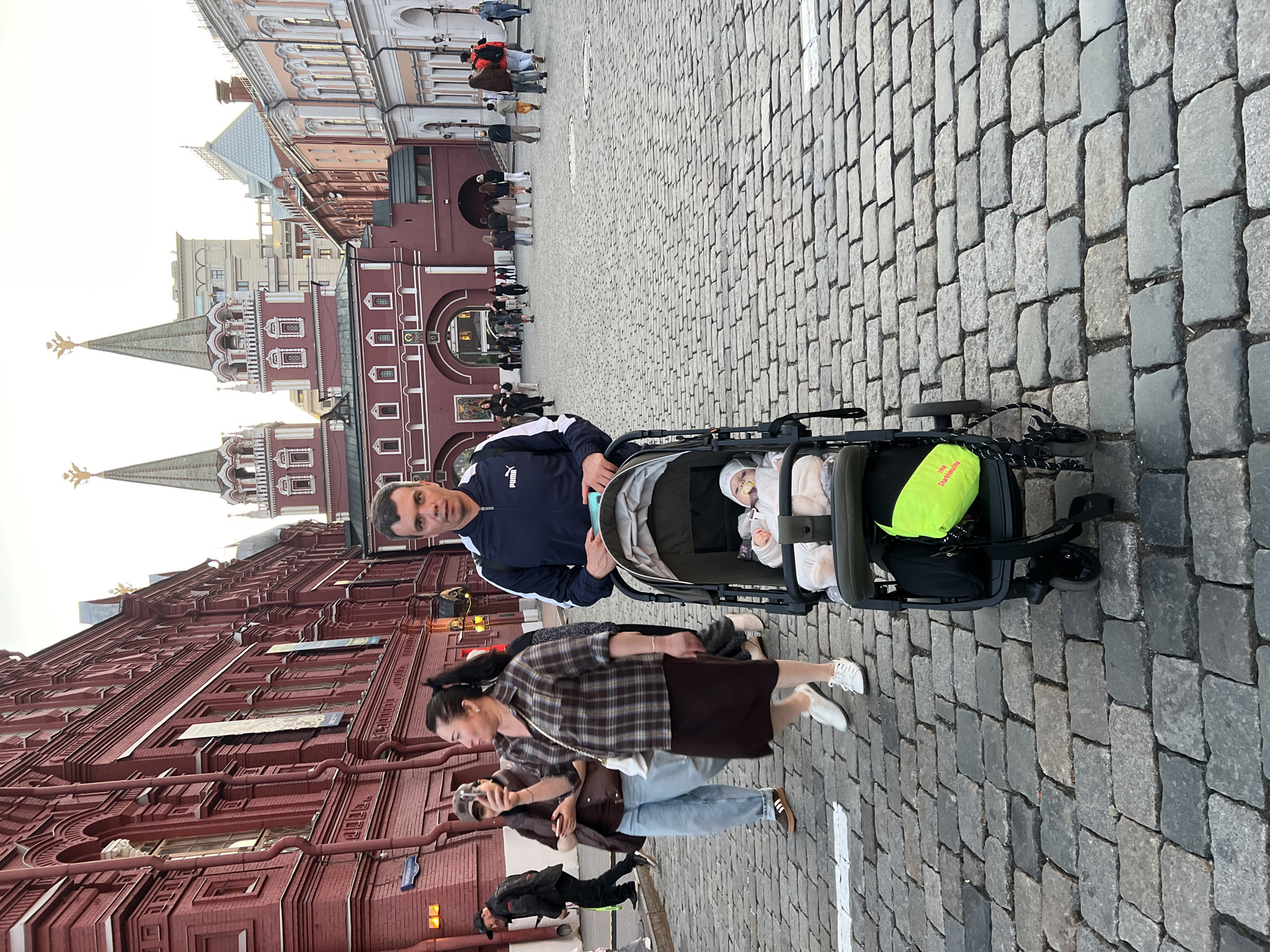 A man stands in the foreground of the image, pushing a baby stroller. The man is wearing a dark blue and white Puma jacket and appears to be looking at the camera. Inside the stroller, a baby is visible, dressed in a white outfit and wearing a pacifier. A bright neon green bag is attached to the stroller.
In the background, a woman in a brown plaid shirt and dark skirt is walking, holding a phone up as if taking a picture. Another person in a dark jacket and a person in a brown vest and light blue jeans are walking behind her. Several other people are scattered in the background, and a large, ornate red building with distinctive towers dominates the cityscape. The ground is paved with cobblestones. The scene appears to be an outdoor public space, possibly a historic city square.