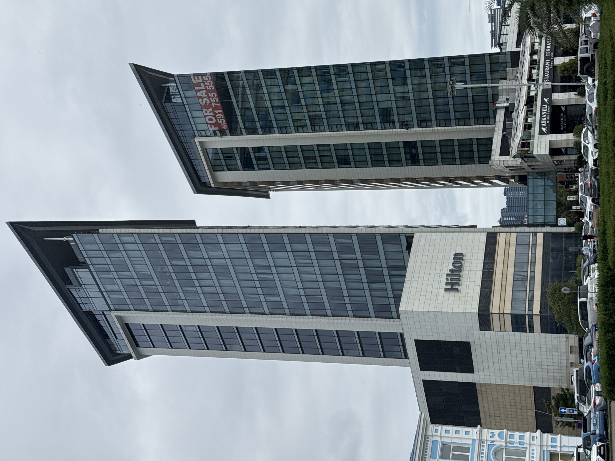 The image shows two modern skyscrapers in Batumi, Georgia. The skyscraper on the right has a red "FOR SALE" banner with a Georgian phone number. The skyscraper on the left has "Hilton" written on its base. Several cars are parked in front of the buildings. The sky is overcast.