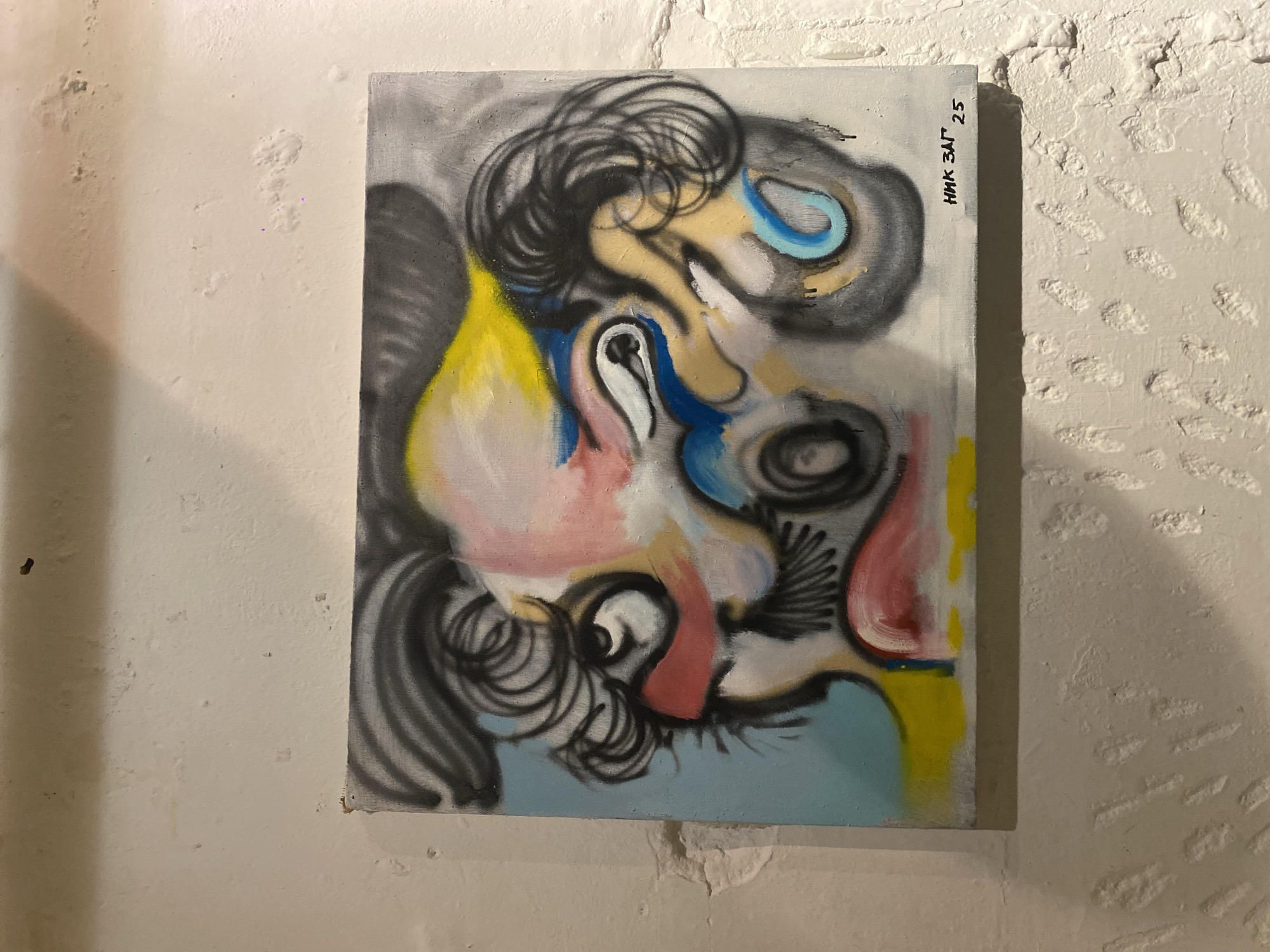 An abstract painting is centrally featured, displayed vertically against a light-colored, textured wall. The artwork itself is characterized by a dynamic interplay of swirling black lines and abstract forms, interspersed with blocks and smudges of color including yellow, blue, pink, and white. The composition hints at fragmented or distorted facial features, such as multiple eyes and sections resembling mouths and noses, all integrated with prominent curly lines that evoke hair. The style suggests a blend of spray paint and possibly other media, creating a vibrant yet somewhat chaotic visual experience.
The painting is mounted on a rough, off-white wall that shows uneven textures and small bumps, indicative of plaster or concrete. A distinct shadow cast by the canvas onto the wall suggests an indoor setting with artificial lighting. No activity is taking place other than the display of the artwork.
A key detail is the vertical inscription on the top right edge of the canvas. Written in black, it reads "НИК ЗАГ 25." This is likely the artist's signature, "NIK ZAG," followed by a number, potentially indicating the year (e.g., 2025) or an edition number. The overall scene depicts a piece of contemporary abstract art within what appears to be a gallery, studio, or exhibition space in Moscow, Russia.