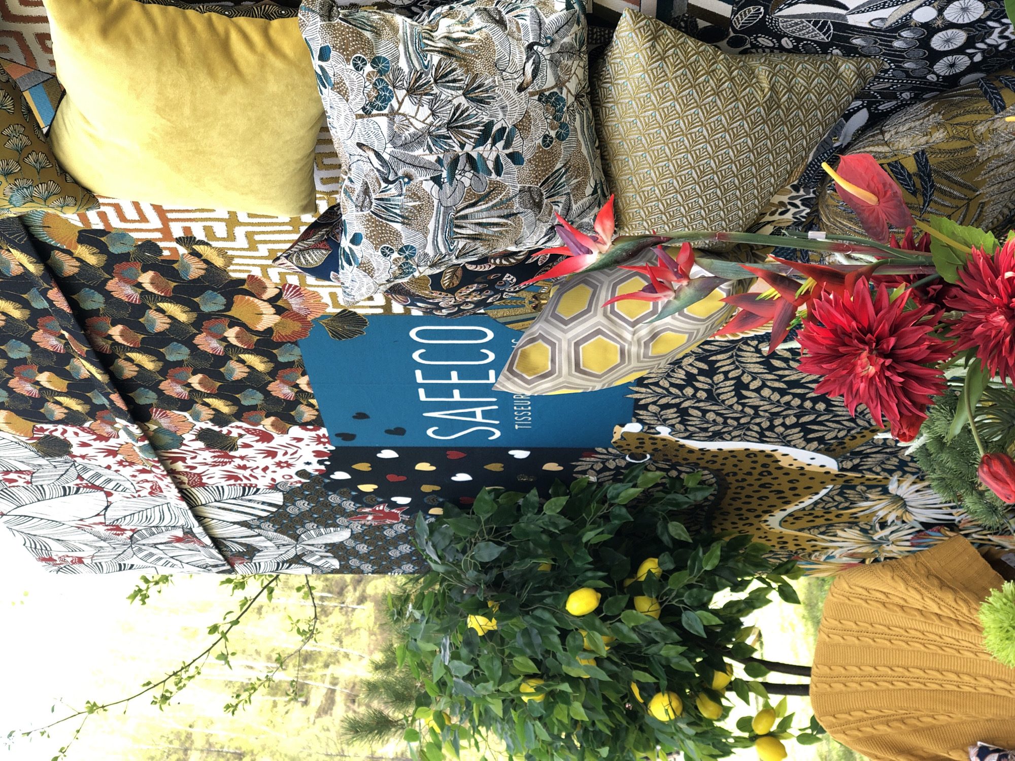 The image shows a collection of cushions and fabrics arranged together indoors, along with floral and plant arrangements. There are several decorative pillows with varied patterns and textures, including a solid yellow velvet cushion, cushions with botanical prints, geometric patterns, and a mustard-colored cable knit pillow. A book or sample card with the text "SAFECO" and "TISSEUR" is visible in the center of the arrangement.
Floral arrangements with red dahlias and other tropical flowers are situated on the right. A lemon tree with yellow lemons is placed on the lower part of the image. The background shows a glimpse of greenery and light suggesting a window or outdoor setting.
The image does not show or imply anything about the city of Krasnoyarsk, Russia.