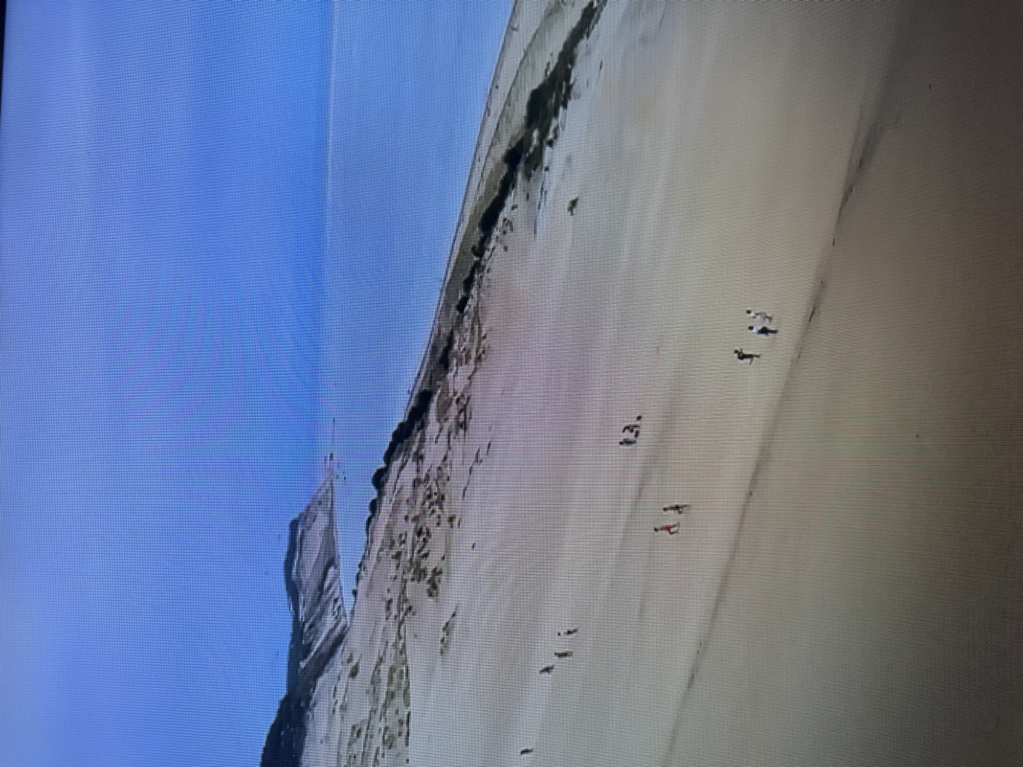 The image depicts a vast expanse of sand dunes meeting the ocean under a clear sky. Several individuals, appearing as small figures from this aerial perspective, are scattered across the sand. Some are walking, while others appear to be standing. The landscape is characterized by the rolling curves of the sand dunes, with sparse vegetation in some areas. The ocean is visible as a calm, blue expanse adjacent to the dunes. The scene is peaceful and expansive, with the focus on the natural landscape and the distant human presence. There is no identifiable information in the image that would place it in the city of Kyiv, Ukraine.