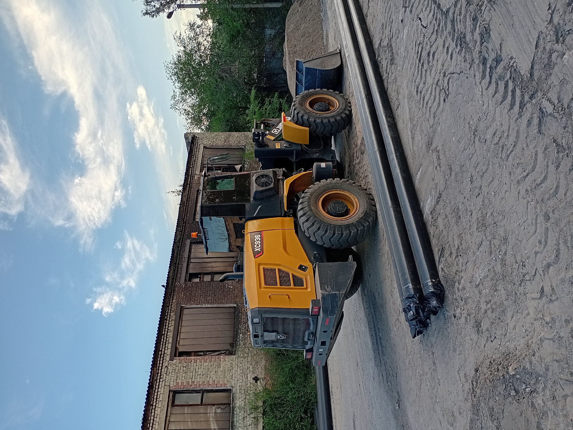 An industrial scene unfolds under a partially cloudy sky in Semey, Kazakhstan. A yellow and black front-end loader, identified by the "XC936" marking, is parked on a dirt and gravel surface. In front of the loader, several large black pipes are laid out, bundled together at one end with black plastic wrap.
Behind the loader, a weathered brick building with large, closed wooden doors and broken windows stands. The building's roof appears to be corrugated metal. Green bushes grow along the side of the building. To the right of the loader, a pile of sand is visible, and a street lamp stands further in the background. The overall impression is one of an active construction or industrial site. There are no people visible in the image.