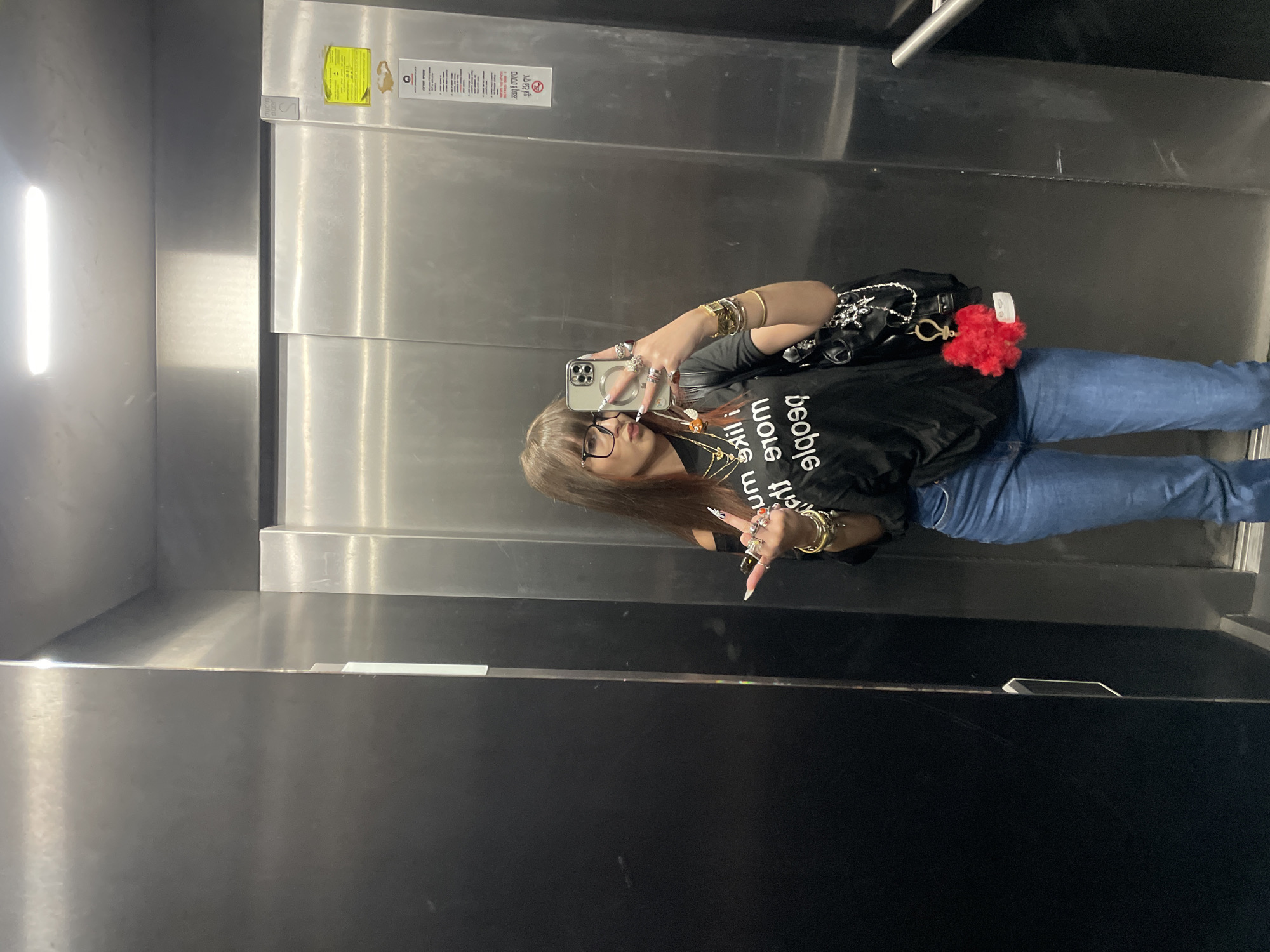 The image shows a woman taking a selfie in an elevator. The elevator appears to be in a commercial or residential building based on the metal walls and ceiling. The woman is wearing jeans, a black shirt, and jewelry. She is holding a smartphone in her hand. There are signs posted inside the elevator, typical of safety or informational notices. Based on the prompt specifying the location in Raanana, Israel, it can be concluded the scene occurs in Raanana, Israel; however, the image provides no information about this location.
