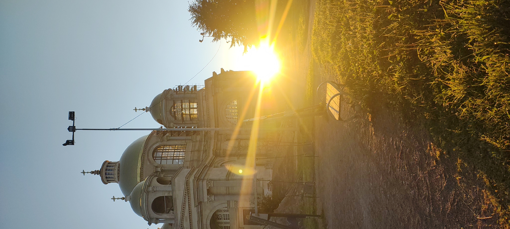 The image shows a side view of a large, ornate building with golden domes, possibly a cathedral or a church, bathed in the bright, golden light of a setting or rising sun. The building's architecture features classical columns, arched windows, and smaller domed structures. Scaffolding is visible on some parts of the building, suggesting ongoing construction or restoration. In the foreground, there is a dirt path and a patch of green grass. A park bench sits on the edge of the path, illuminated by the sun's rays. A playground swing set is partially visible behind the bench. The sun's glare is intense, creating lens flare effects across the image. The sky is a clear, pale blue. A tall light pole with multiple lamps stands near the building. The overall scene evokes a sense of tranquility and historical grandeur, possibly captured in the late afternoon or early morning. This building is likely the Smolny Cathedral, located in the city of Saint Petersburg, Russia.
