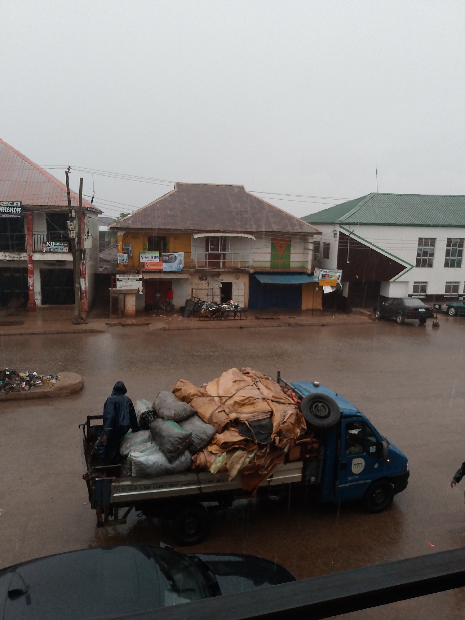 The image depicts a street scene in Bukuru, Nigeria, during heavy rainfall. The street is partially flooded. A small blue truck is heavily laden with bags and a spare tire, partially covered by a tan tarp. A person in a dark hooded raincoat is standing on the back of the truck. In the background, buildings line the street, including a two-story structure with a yellow facade and advertisements for "The SIM." Additional cars are parked along the street. A pile of garbage is visible near a traffic island in the foreground. The sky is overcast and grey, reflecting the rainy conditions.