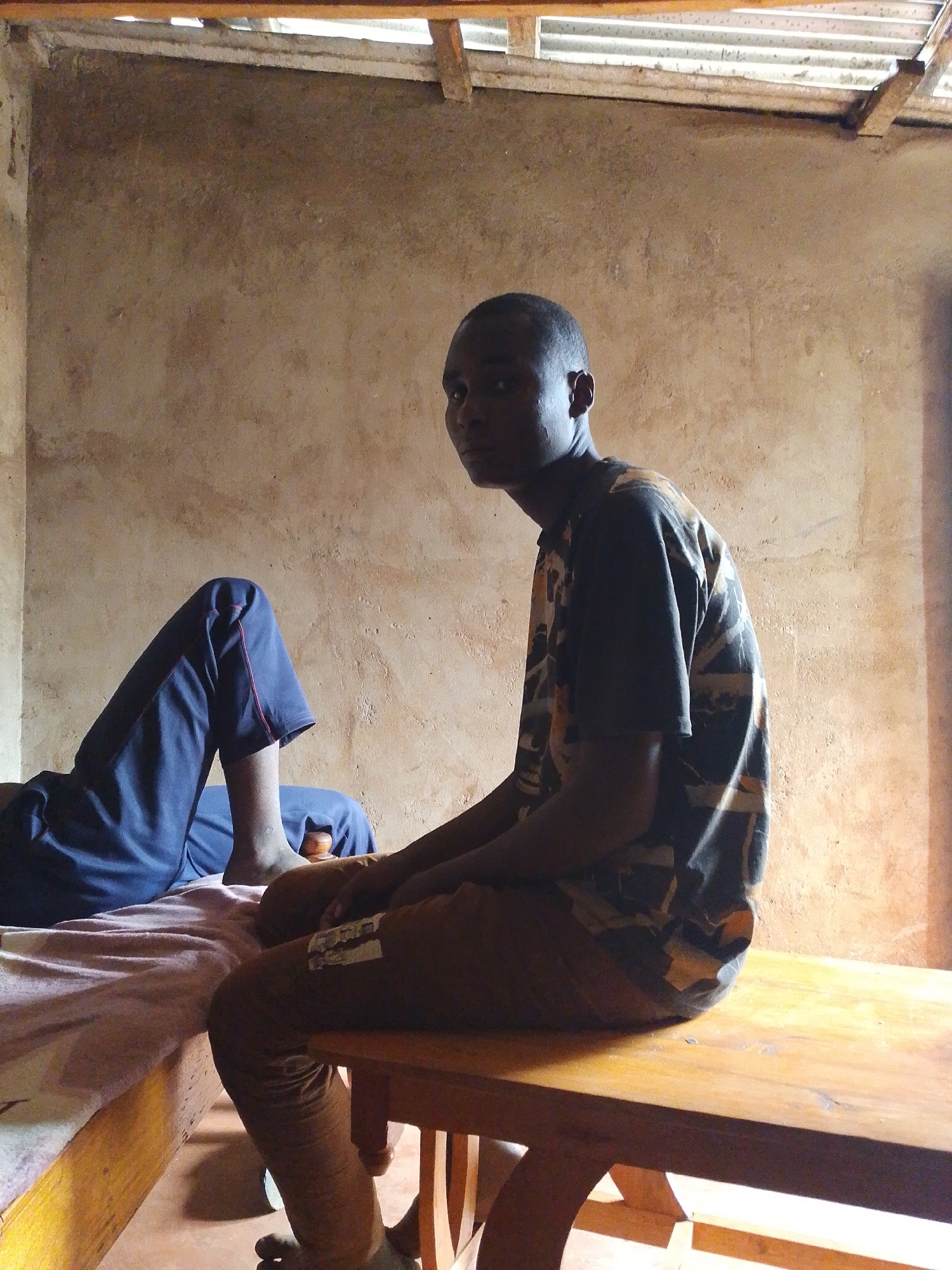 A young male, with dark skin and short hair, is seated on a wooden bench in a room with rough, textured walls and a corrugated metal ceiling supported by wooden beams. He is wearing a short-sleeved shirt with a dark camouflage pattern and brown trousers. To his left, another person, mostly obscured, is lying down on a bed, wearing dark blue trousers. The room appears to be sparsely furnished and dimly lit, with light streaming in from an unseen source. The overall impression is of a simple, domestic setting within the Borabu Chitago ward, Kenya.