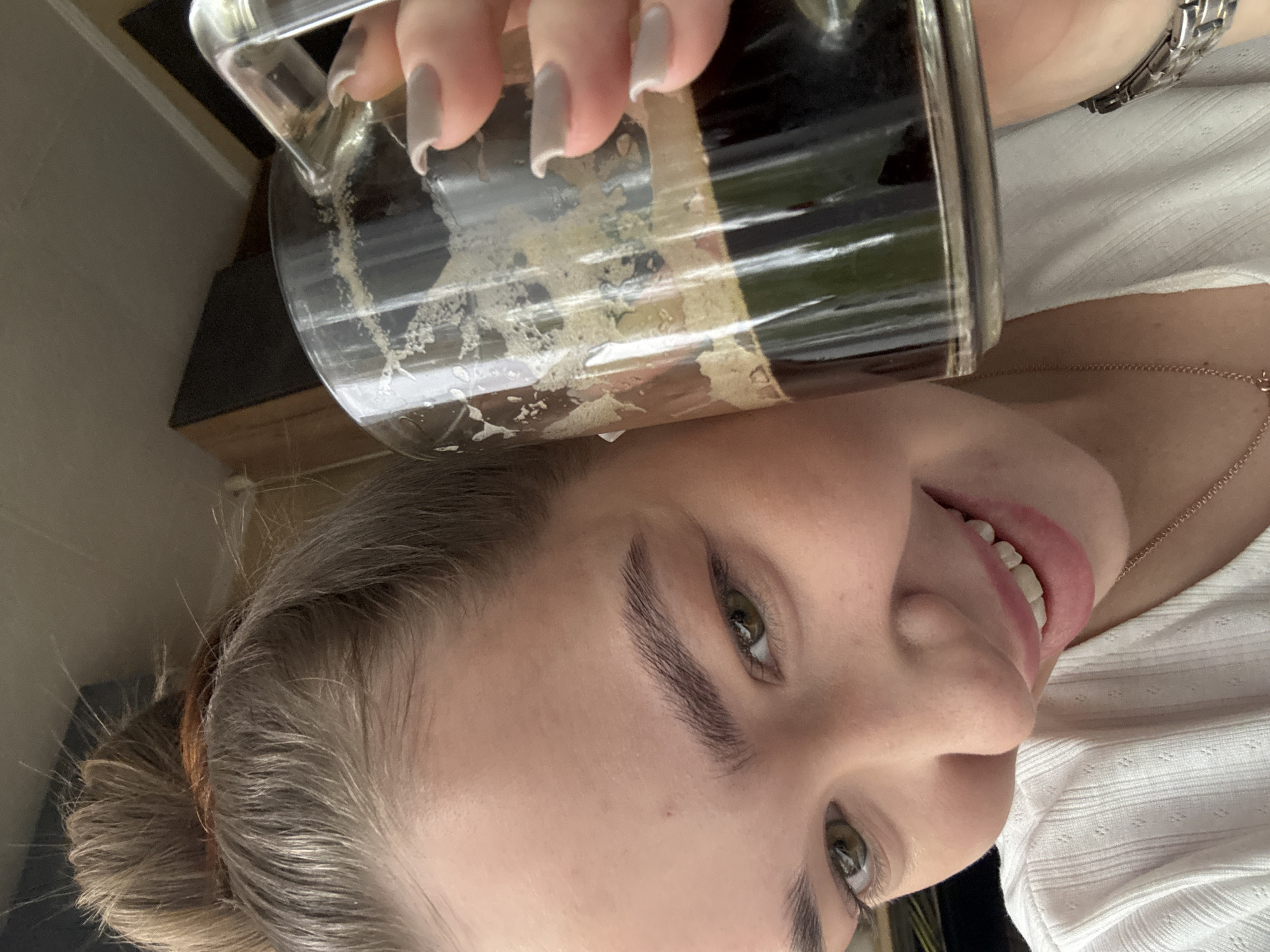 A woman holds a glass mug filled with a dark liquid, likely coffee or stout, with foam clinging to the glass. She is wearing a white, textured shirt and a watch on her wrist. Her hair is pulled back in a bun, and she has light-colored fingernails. A gold necklace is visible around her neck. The background is blurred, but appears to be an indoor setting with a wooden shelf or table. The location in Chaikovsky, Russia, cannot be determined from the image.