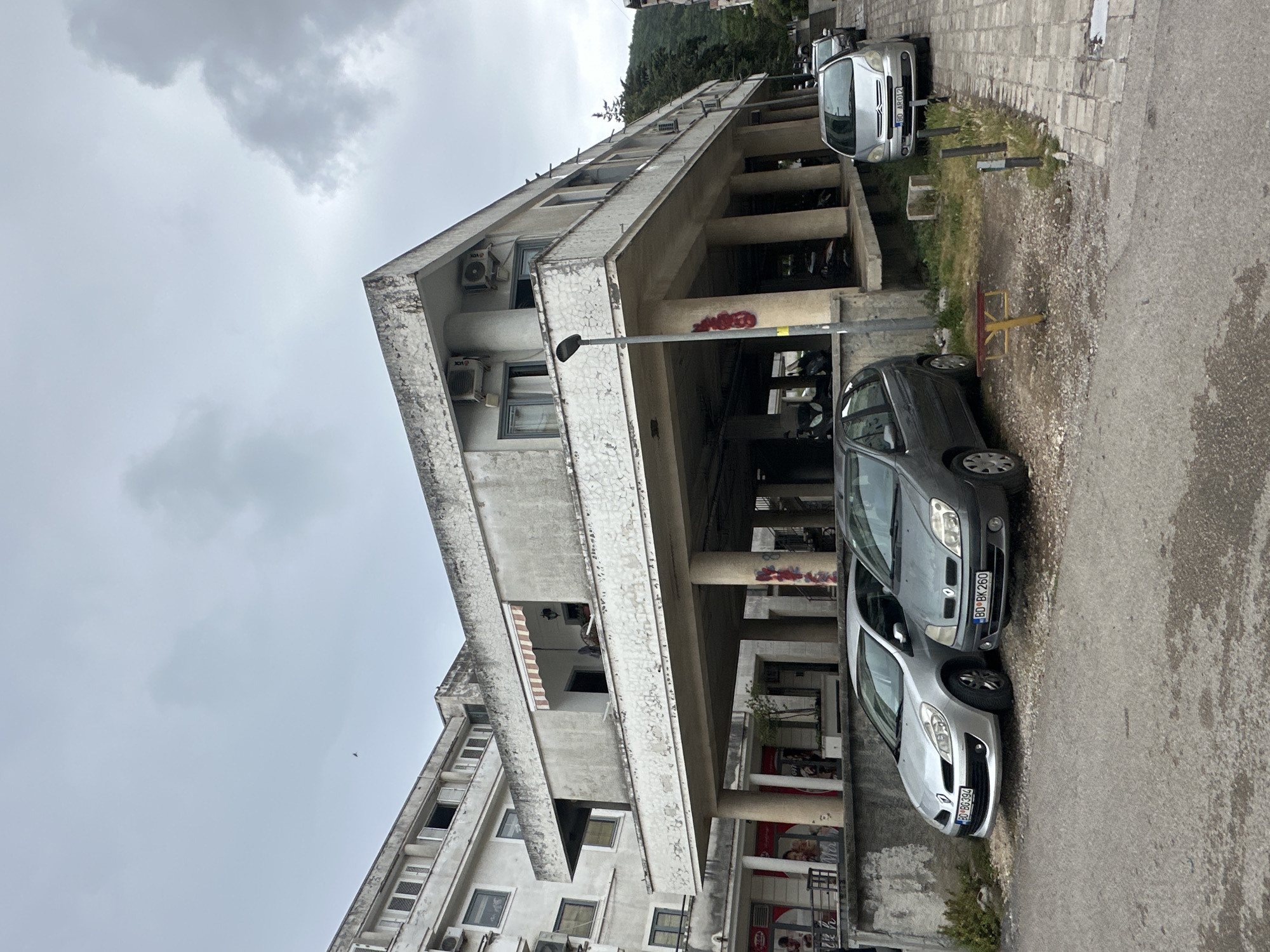 The image shows a concrete building in Budva, Montenegro. The building has a distinct architectural style, featuring columns that support the upper floors, creating a covered walkway or parking area at ground level. There are several cars parked on the street adjacent to the building; a silver Citroen C3 Picasso with license plate 'BD 80-AB012', a grey Renault Clio with license plate 'BD BK-260', and a silver Renault Clio with license plate 'BD BG394'. Graffiti is visible on some of the columns supporting the building. Air conditioning units are mounted on the exterior walls of the building. The sky is overcast. A street lamp is in front of the building.