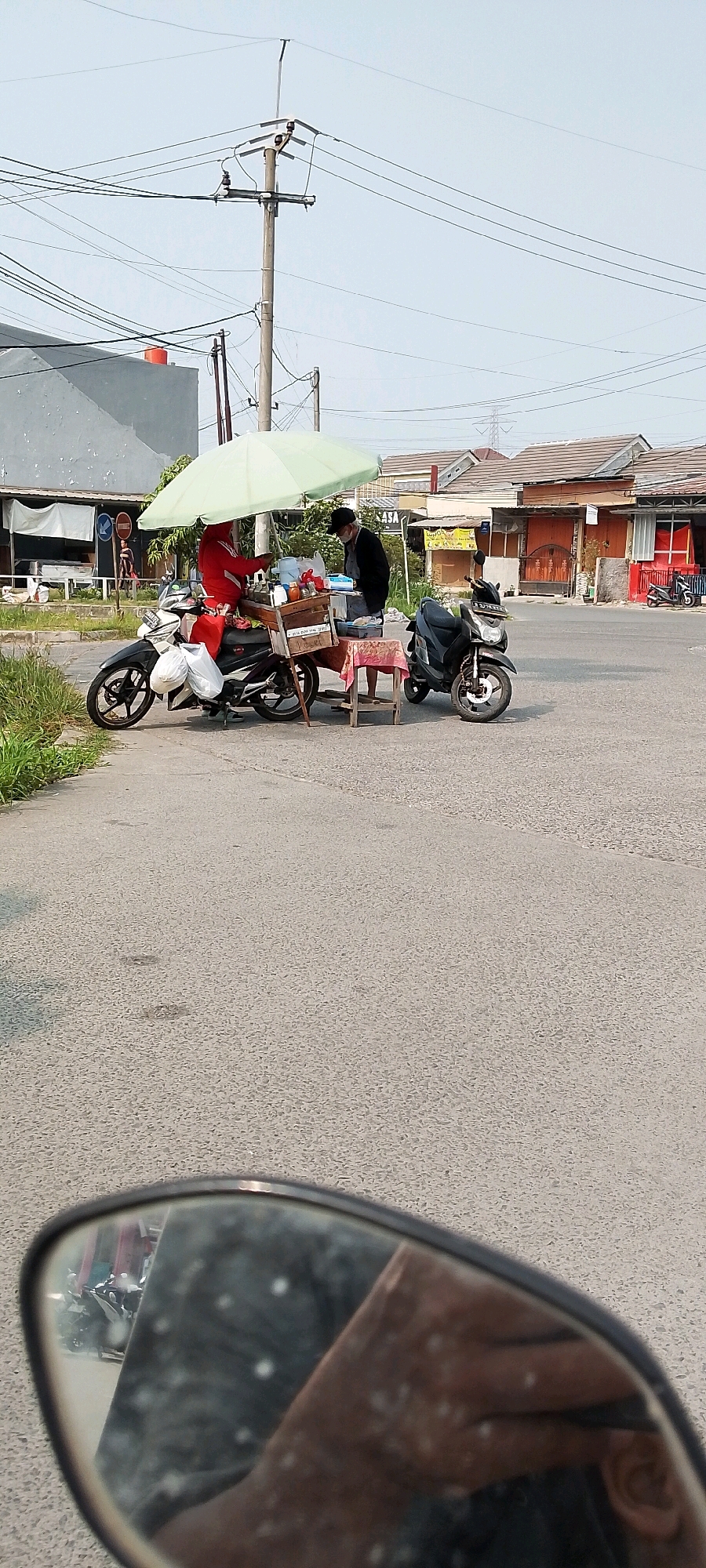 In Satriamekar, Indonesia, on a paved road, two individuals are engaged in commerce from a mobile cart situated on the back of a motorcycle. A person wearing a red jacket and a head covering is seated on the motorcycle, managing the cart. Another individual, wearing a dark jacket and a face mask, stands beside the cart, appearing to interact with the vendor or customer. The cart itself is a wooden structure attached to the motorcycle, laden with various items, including bottles and containers, and is shaded by a large, light green umbrella. A second, dark-colored motorcycle is parked nearby. The background shows residential buildings with tiled roofs and utility poles with numerous power lines crisscrossing the sky, indicating an urban or suburban setting. The scene is captured from a low angle, possibly from the perspective of another vehicle, with a motorcycle's side mirror visible in the foreground reflecting a portion of the scene and the photographer.