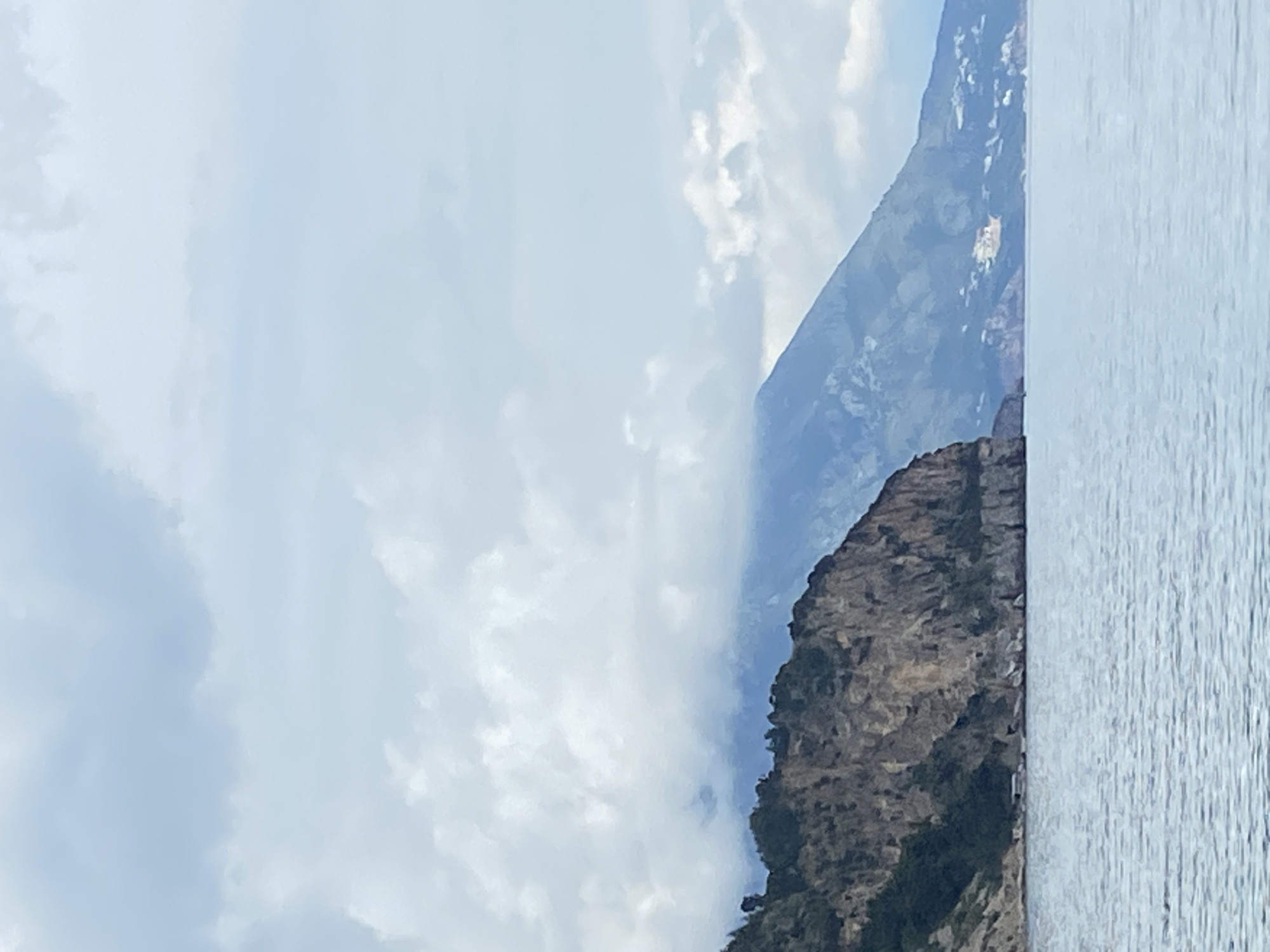 The image depicts a cliffside along the coast of Prijevor, Montenegro. The cliff face, which is predominantly gray and rocky, is partially covered in vegetation. To the right of the cliff is a body of water with a light blue tint. Above the cliff and water is a sky filled with light, hazy clouds. The image is oriented vertically.