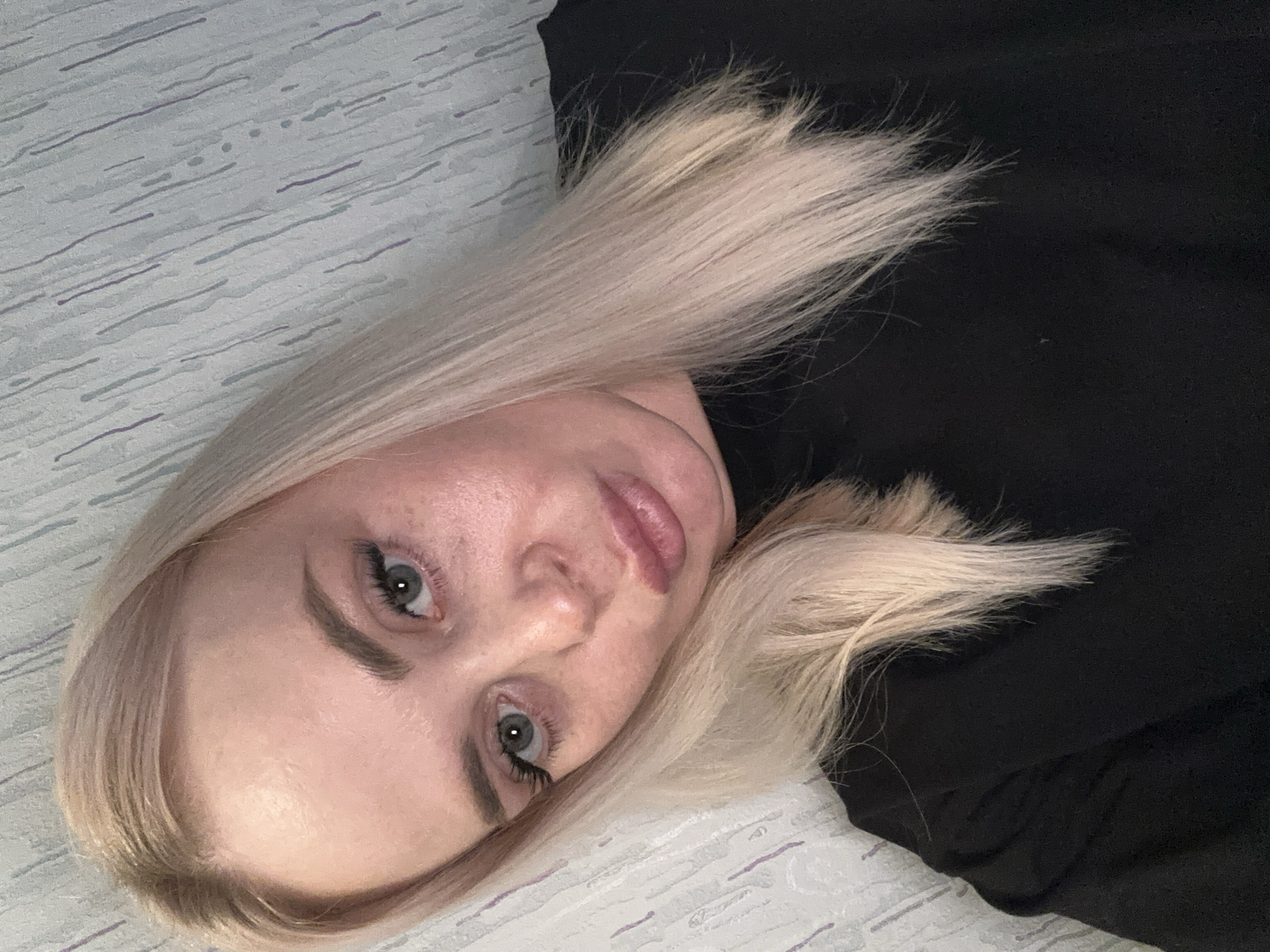 A young Caucasian woman with blonde hair and blue eyes is shown in a close-up, angled selfie. She is wearing a black top and is positioned against a backdrop of light blue wallpaper with a subtle pattern of vertical lines and dots in shades of gray and purple. Her hair is parted on the side and falls loosely around her face and shoulders. Her expression is a pout, with her lips slightly parted. The lighting appears to be natural, casting soft shadows on her face. The image quality is good, with clear details of her features and the background. The scene is indoors, likely in a residential setting.