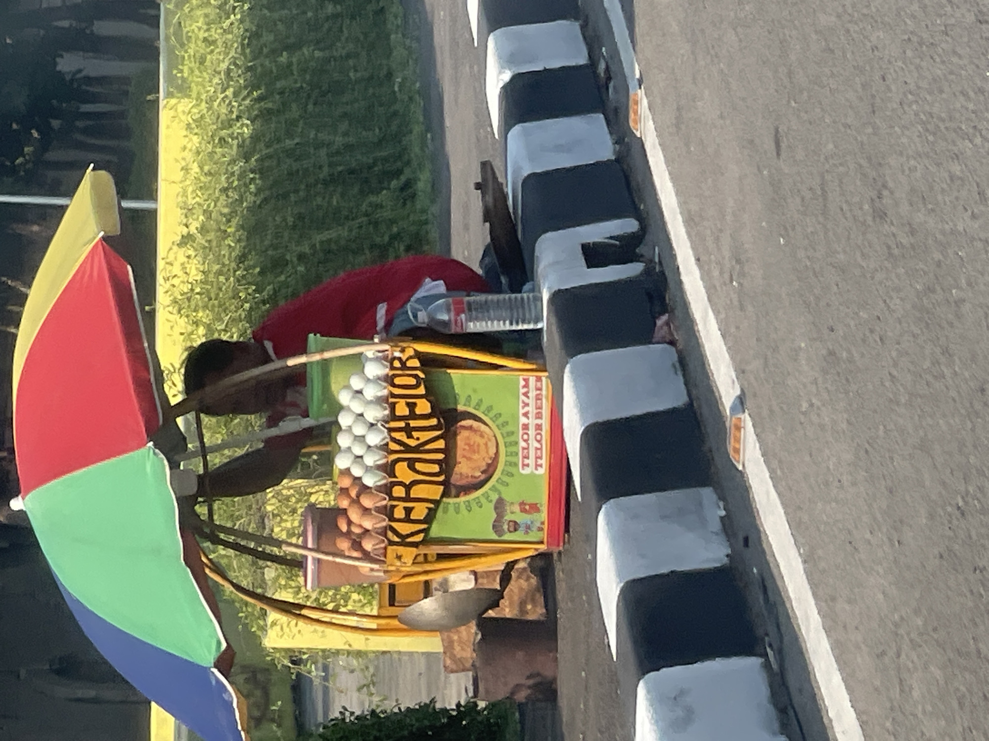 The image depicts a street vendor selling "Kerak Telor," a traditional Indonesian omelet, from a cart under a colorful umbrella. The cart is positioned alongside a road with a black and white striped concrete barrier. A person, presumably the vendor, is seated behind the cart, wearing a red shirt. The cart itself is yellow with a green front panel that displays "KERAK TELOR" in yellow lettering. Below this, there are illustrations of two chefs and text indicating "TELOR AYAM TELOR BEBEK" (chicken eggs and duck eggs). The cart holds a variety of items, including eggs in two separate containers (brown eggs and white eggs) and a bottle of water. To the left of the cart, there's a large, dark metal pot. The background shows lush green foliage and trees. This scene is located on a roadside in North Jakarta, Indonesia, as indicated by the street furniture and general urban environment characteristic of the area.