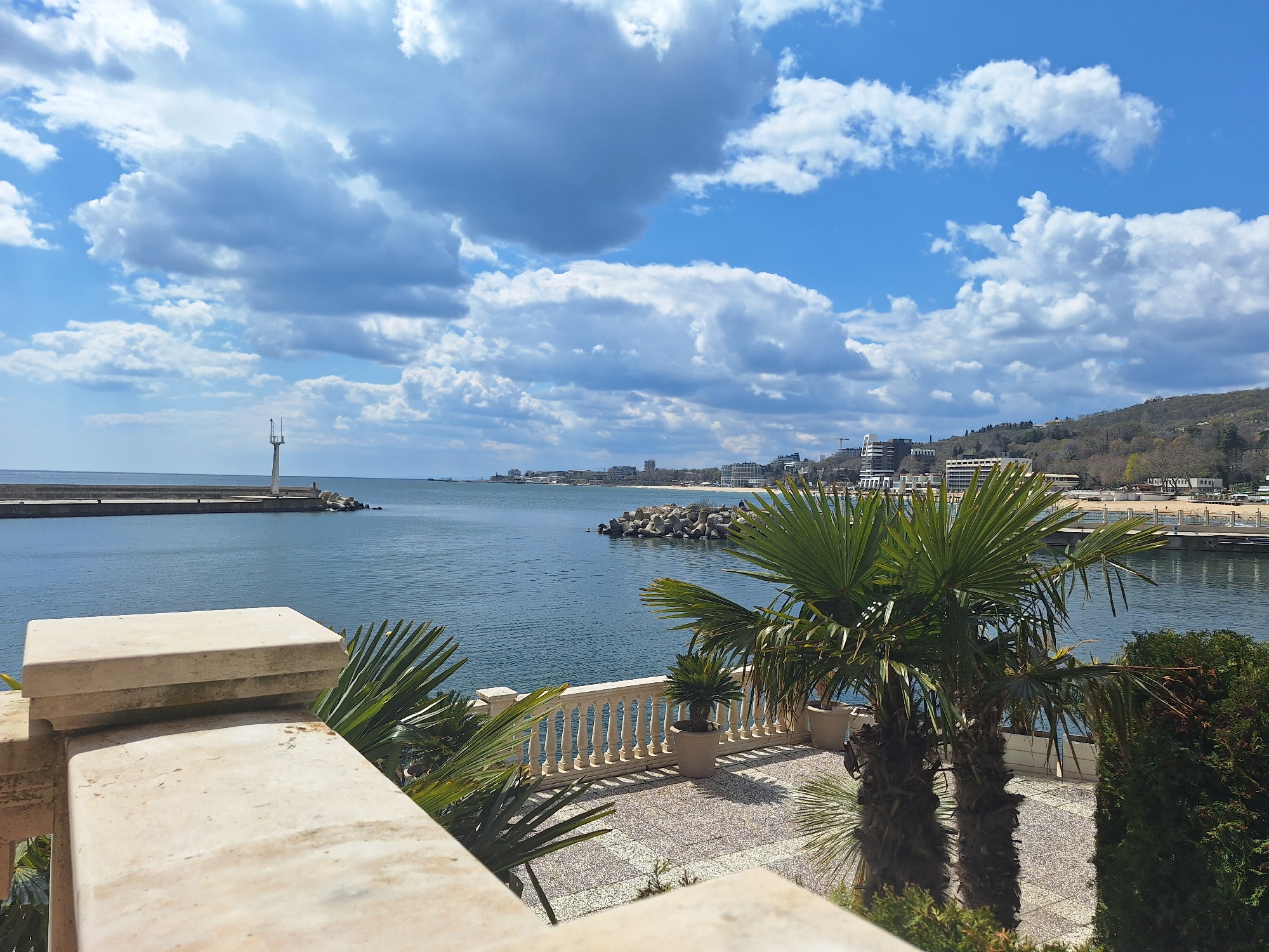 View from a balcony overlooking the Black Sea in Varna, Bulgaria. The sky is blue with cumulus clouds. In the distance, a lighthouse stands at the end of a concrete pier. Along the coast, buildings and trees are visible. The balcony has a white stone balustrade and a tiled floor. Palm trees grow in pots and in the garden. Green bushes surround the balcony.