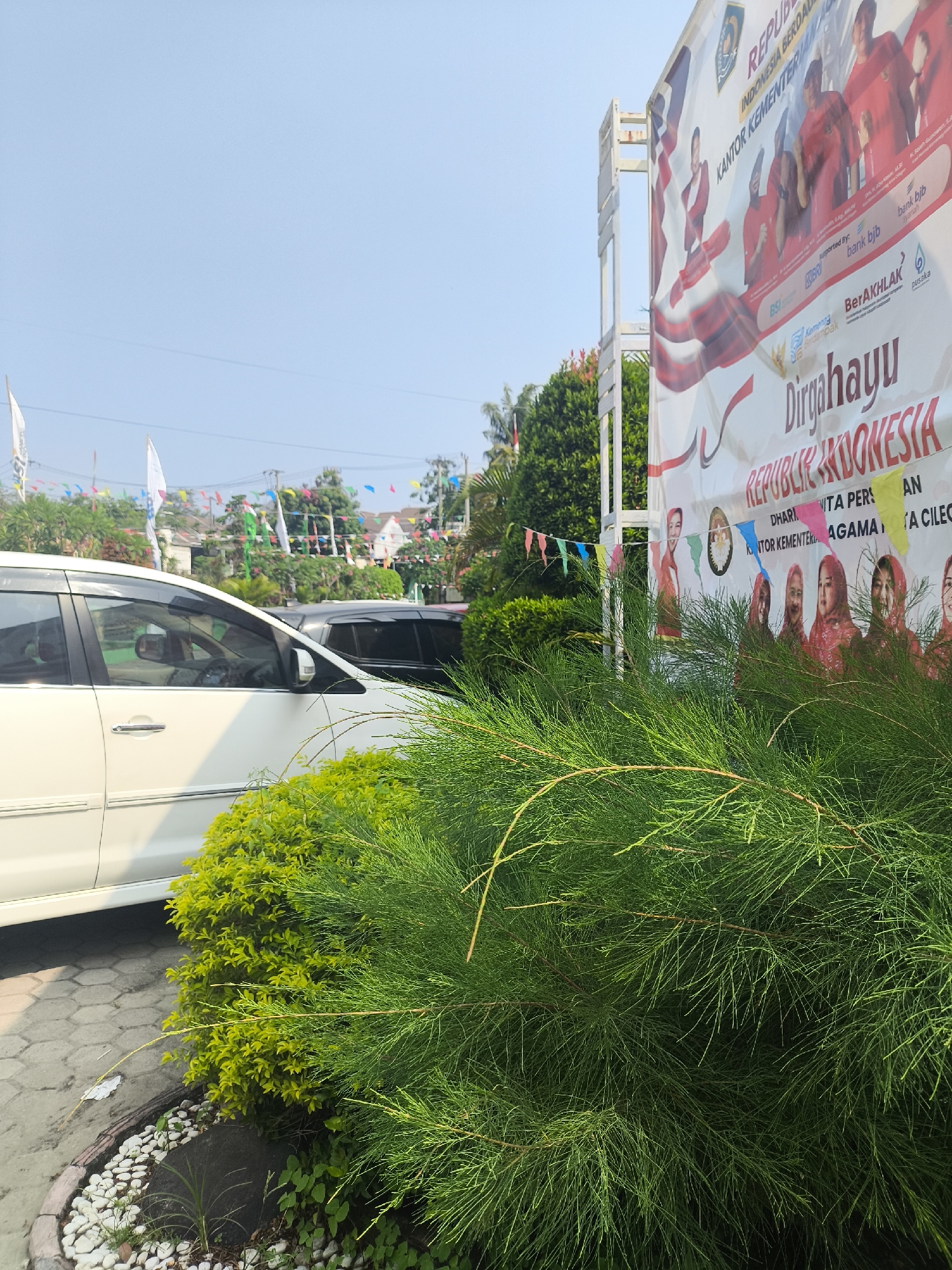 A large banner displaying "Dirgahayu Republik Indonesia" and a portion of the Indonesian flag is prominently featured on the right side of the image. Below the text, there are several images of women in red headscarves. The banner is partially obscured by a dense green shrub in the foreground.
To the left of the banner, a white car is parked. Behind the car, another dark-colored car is visible. Further in the background, there are trees, buildings, and several small, colorful flags strung across a wire, suggesting a festive occasion. The sky is clear and blue. The overall scene appears to be an outdoor setting, possibly a public space or a street in the city of Cilegon, Indonesia, given the presence of the banner referencing Indonesian Independence Day and the mention of "Cilegon" in the banner text. The ground in front of the car and shrub is paved with hexagonal concrete tiles and decorated with small white stones and a larger dark stone.