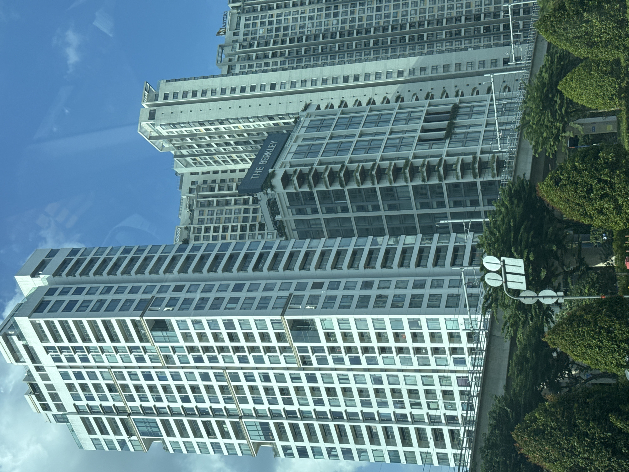 The image captures a view of modern high-rise buildings in the city of Thủ Đức, Vietnam, likely taken from a moving vehicle. In the foreground, lush green trees and a traffic signal with three circular lights and two rectangular lights are visible. A concrete overpass stretches across the mid-ground, with white railings. The background is dominated by several contemporary apartment buildings. The tallest building on the left is predominantly white with numerous balconies and windows. To its right, another tall building features balconies and dark-tinted windows. Further to the right, a lighter-colored building displays rows of windows and has "MASTERY" signage visible near its top. A building labeled "THE BERKLEY" with a dark facade and green vertical gardens on its balconies is positioned in front of the taller buildings. The sky is blue with scattered white clouds.