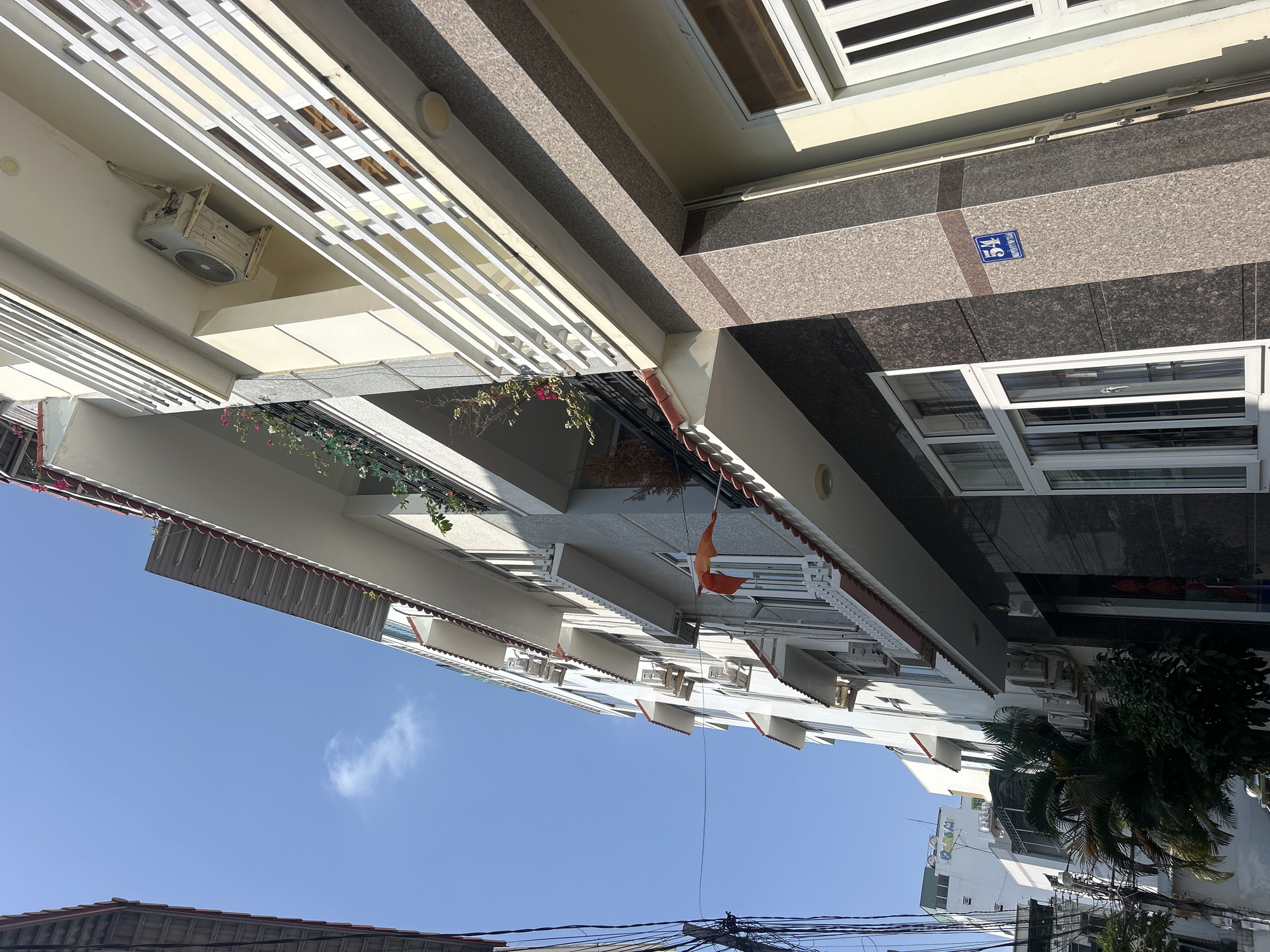 The image depicts an upward-angled view of buildings and the sky in Bắc Nha Trang, Vietnam. The facades of multi-story buildings dominate the scene. Notable details include air conditioning units mounted on exterior walls, balconies with floral arrangements, and a Vietnamese flag. A building displaying the number 15 is also visible. Electrical wires are strung across the lower portion of the frame, while parts of rooftops and a palm tree are seen in the lower part of the image. The sky is a clear blue with scattered cloud formations.