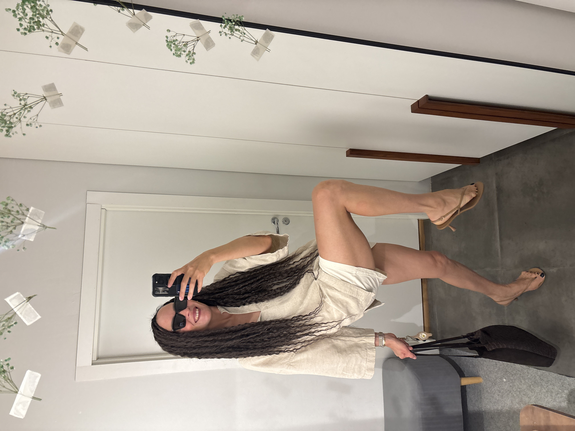A woman with long, dark braided hair, wearing sunglasses, is captured in a mirror selfie indoors. She is smiling broadly and holding a black smartphone with a prominent camera in her right hand to take the photo. Her left hand holds a black handbag with two straps, and a silver-toned watch is visible on her left wrist. She is dressed in a light-colored, possibly linen, short-sleeved button-up top and matching shorts. Her legs are visible, with her feet clad in light-colored heeled sandals, showing dark nail polish on her toes.
The setting appears to be a modern interior space, possibly a dressing room or studio. The primary background is a large mirror reflecting the woman and parts of the room. Several small sprigs of green artificial plants are taped to a white surface that is reflected in the mirror, positioned above the woman's head. The mirror has a thin black frame at the top. White walls are visible, along with a white door frame behind the woman. To her right, there are two parallel dark wooden decorative elements or shelves mounted on a white wall. The floor is a dark gray. A piece of gray upholstered furniture, possibly a bench or seat with a wooden accent, is partially visible in the lower right portion of the image. The original image is oriented 90 degrees counter-clockwise.