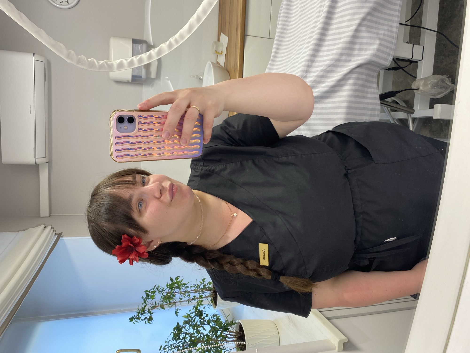 A woman is taking a selfie in what appears to be a beauty salon or clinic. She is wearing a black uniform with a name tag that reads "ВНППA" in Cyrillic, which likely indicates her name or initials. A red flower is tucked behind her ear, and she has a braid in her hair. She is holding a smartphone with a colorful, wavy patterned case to take the picture.
The environment includes an air conditioning unit, a clock, a wall-mounted dispenser, and a treatment bed covered in striped fabric. A potted plant is visible near a window, suggesting natural light. The details provided do not provide enough information to confirm if the location is in Votkinsk, Russia.
