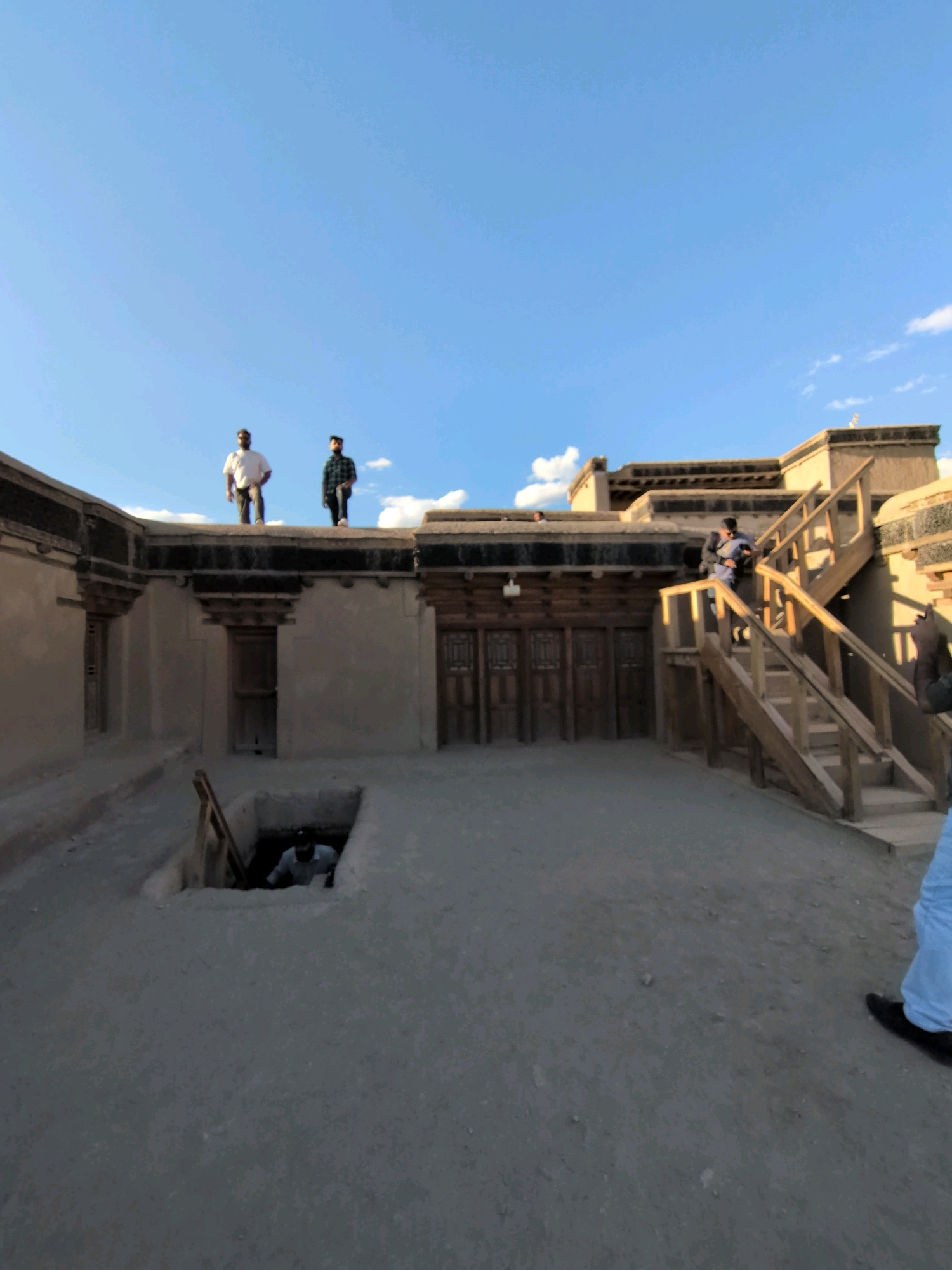 The image captures an outdoor scene at an ancient structure, possibly in Leh, India, characterized by its adobe-like walls and traditional architectural elements. The sky above is a clear blue with a few wispy clouds.
In the foreground, a courtyard paved with rough, grey material dominates the lower half of the frame. To the left, there is a square-shaped opening in the ground, revealing a lower level where a person wearing a light-colored shirt and a cap is visible, possibly descending or standing within the pit. A wooden ramp leads down into this opening.
The mid-ground features the main structure, with thick, unadorned walls. Multiple wooden doors with intricate carved panels are visible along the facade. Above the doors, exposed wooden beams and decorative eaves are apparent. A wooden staircase, weathered and aged, rises on the right side of the structure, leading to an upper level. Several people are present on the upper levels and on the roof.
On the roof of the main structure, two men are standing. The man on the left wears a white shirt and dark pants, while the man on the right is dressed in a dark, plaid shirt and dark pants. They appear to be looking out over the landscape. Further to the right, on a higher section of the building, another man is descending the wooden stairs, carrying a bag over his shoulder. His attire suggests he is wearing a dark jacket and blue pants. On the far right, partially visible in the frame, is a person wearing light blue pants and black shoes, possibly facing away from the camera.
The overall impression is one of historical significance and a preserved architectural site, with visitors exploring its different levels. The lighting suggests it is late afternoon, with shadows cast by the structures and the staircase.