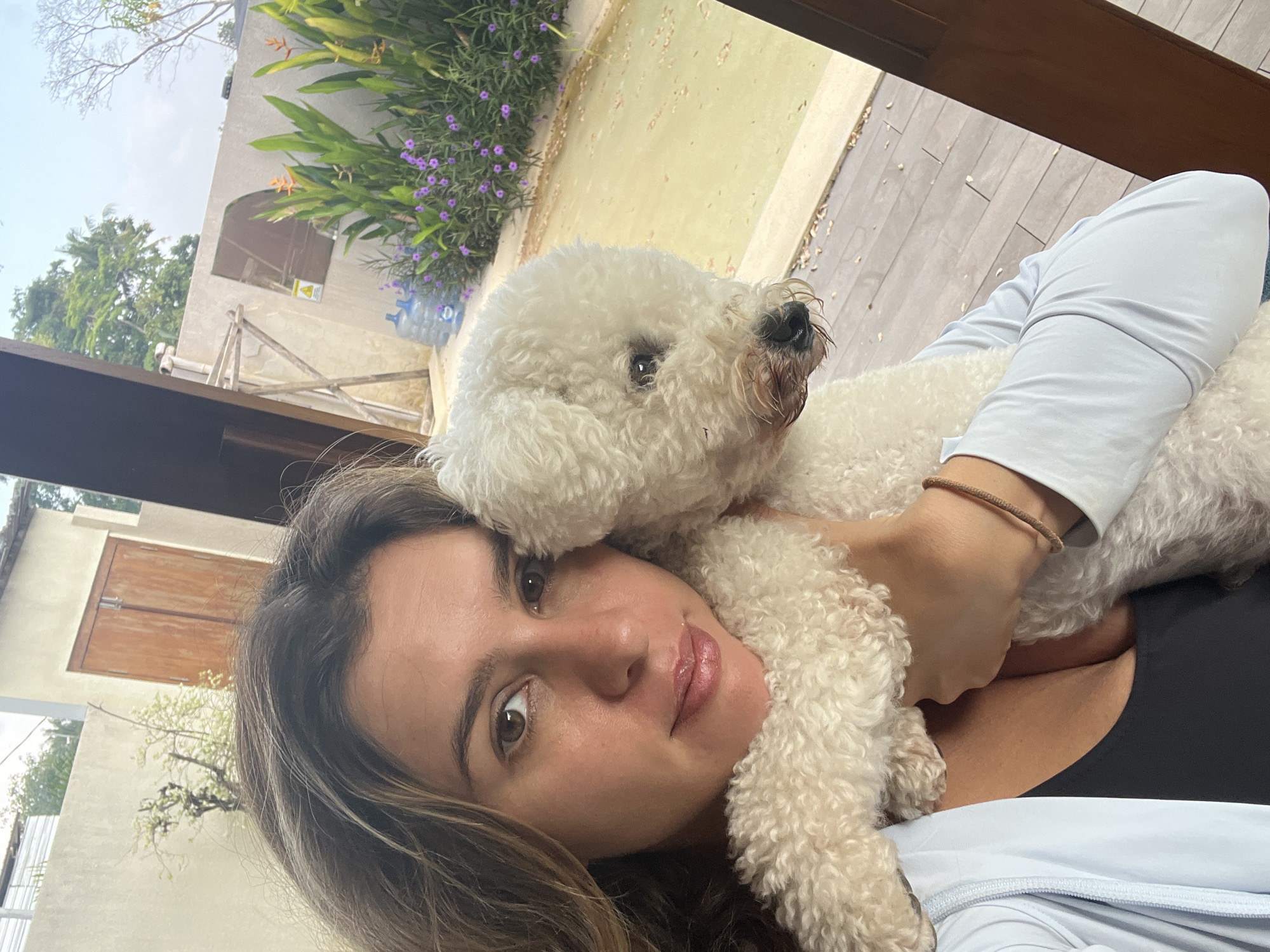 A woman with brown hair and brown eyes, wearing a light blue long-sleeved top over a black sports bra, is taking a selfie outdoors. She is holding a small, fluffy white dog, which is nestled against her chest. The woman's face is turned towards the camera, with the dog partially obscuring her right cheek. In the background, there is a wooden deck, a light-colored wall, and lush green foliage with purple flowers. A large water bottle is visible near the foliage. There are also some wooden doors and scaffolding-like structures in the distance. The overall impression is a casual, outdoor setting, likely in a tropical or subtropical climate. There is no indication that this image is located in the city of Kediri, Indonesia.
