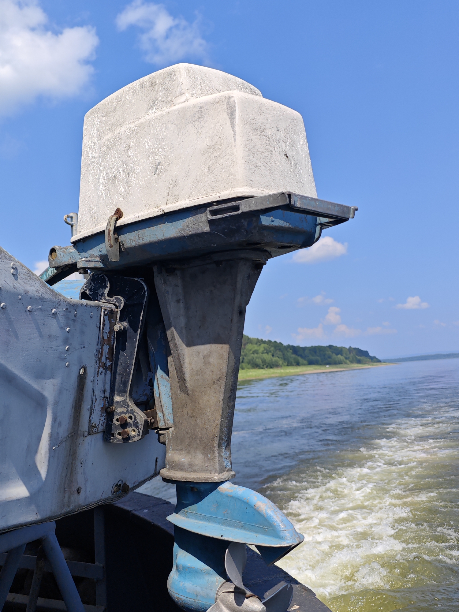 An old, weathered outboard motor is mounted on the stern of a boat, its white hood faded and cracked. The motor is attached to a blue metal bracket that connects to the boat's hull, which is also painted in faded blue and gray. The boat is moving through water, creating a white wake behind it. In the background, a tree-lined shore is visible under a bright blue sky with scattered white clouds. The scene appears to be on a body of water, likely a river or lake, given the calm water and the presence of a shoreline. The location is described as Pogodaevo, Russia, suggesting a rural or semi-rural setting. There are no people visible in the image. The overall impression is one of a functional, albeit aged, piece of marine equipment operating in a natural environment.