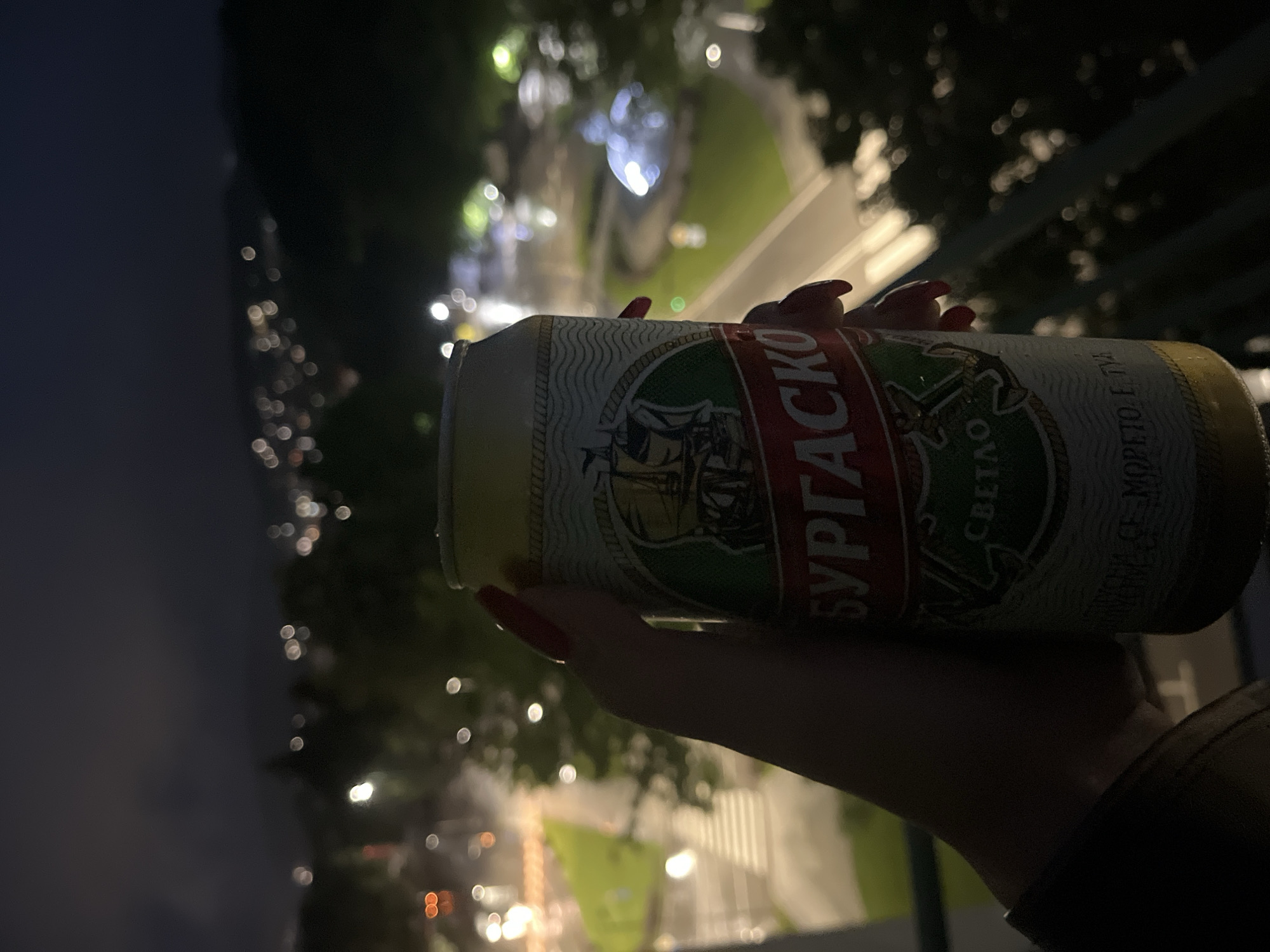 A person's hand, adorned with bright red nail polish, holds a metal beverage can in a casual grip. The hand and can are positioned diagonally across the frame. The can's label is prominent, featuring a green and red design with a stylized sailing ship logo. Visible text on the label includes "БУРГАСКО" (Burgasko), identifying it as a Bulgarian beer, and "СВЕТЛО" (Svetlo), indicating it's a light lager. Another phrase, "МОРЕТО Е ТУК" (The Sea is Here), is also visible, hinting at the brand's coastal association.
The scene is set at night, viewed from an elevated position, possibly a balcony or terrace. The background is a heavily blurred urban or semi-urban landscape, filled with numerous out-of-focus lights creating a bokeh effect. These lights suggest illuminated pathways, buildings, and possibly trees or foliage, with patches of green indicating grass or park areas below. The overall ambiance is dark and relaxed, typical of an evening setting. No specific weather conditions are discernible, but the lights suggest a clear night. The activity depicted is simply holding and likely enjoying a drink while observing the nighttime view.