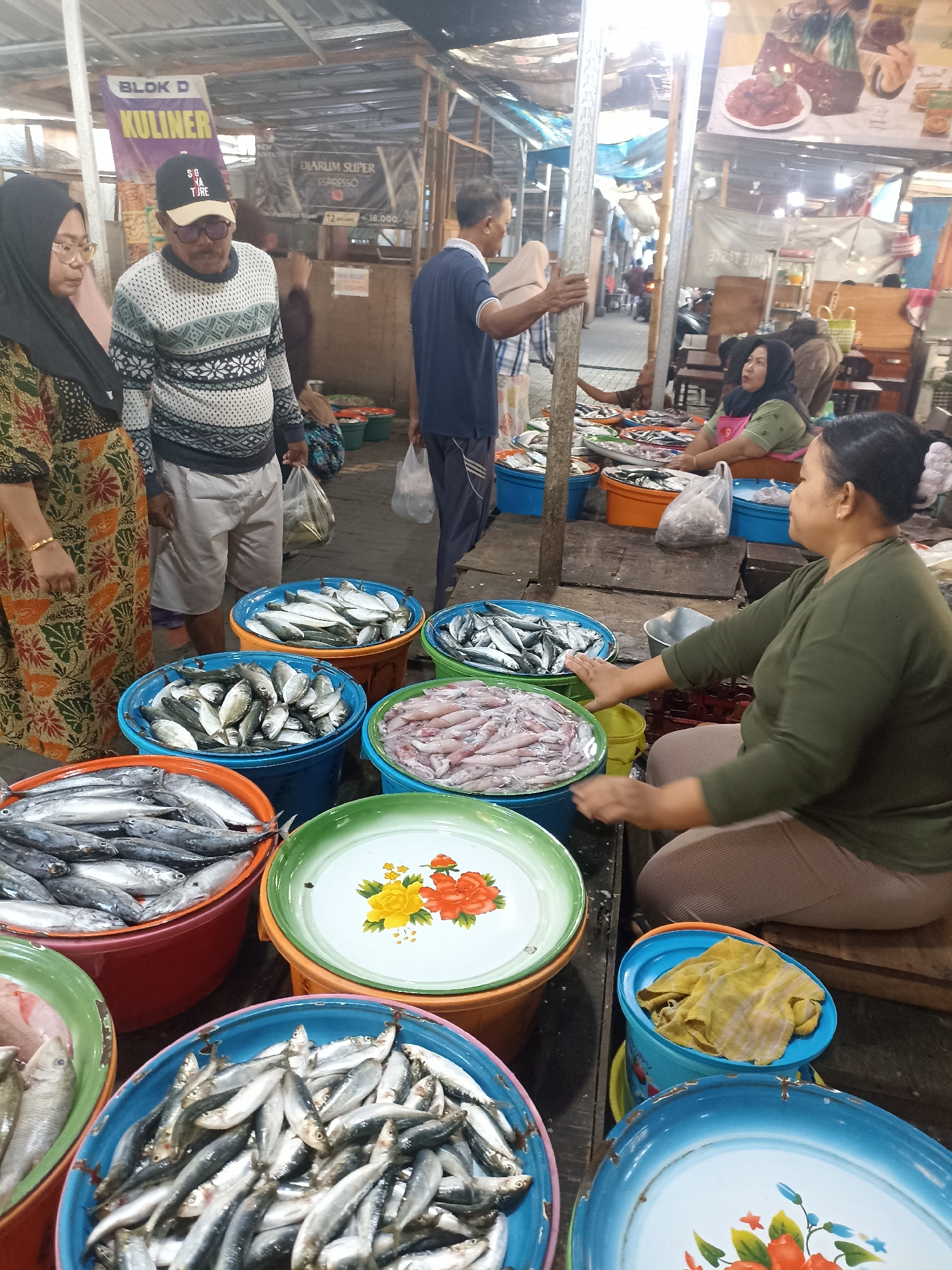 The image depicts a bustling fish market in Kelurahan Kepatihan, Indonesia. In the foreground, several large buckets overflow with various types of fish. A woman in a green shirt and headscarf is seated behind a stall, tending to the fish. To her left, a man in a patterned sweater and a cap is interacting with a woman wearing a hijab and an orange-patterned dress. In the background, other shoppers and vendors are visible, creating a lively atmosphere. The market appears to be covered, with a corrugated metal roof overhead. Signs indicating "BLOK D KULINER" and "DIARUM SUPER ESPRESSO" are present, suggesting a designated culinary or market section. The overall scene captures the daily commerce and vibrant local culture of the area.