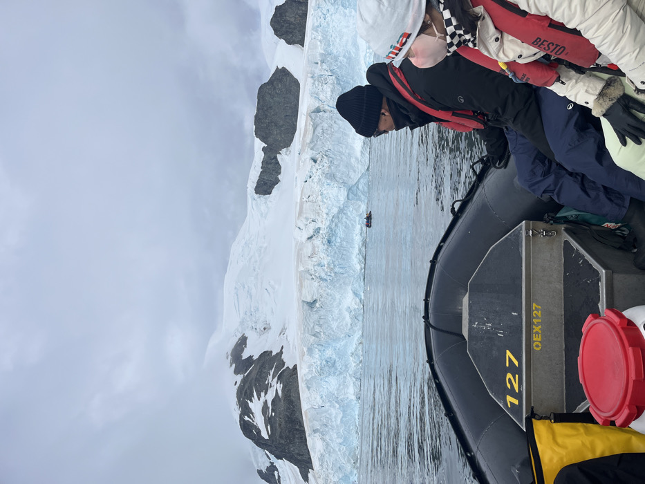 A group of people in an inflatable boat observe a large, white glacier with rocky peaks on a cloudy day. One person in the boat is leaning forward, looking at the water. The boat's side shows the number "127" and "QEX127" on a container. There is another smaller boat floating along the glacier. The clothing worn by the people in the boat includes winter hats, life jackets, and parkas, indicating a cold environment.