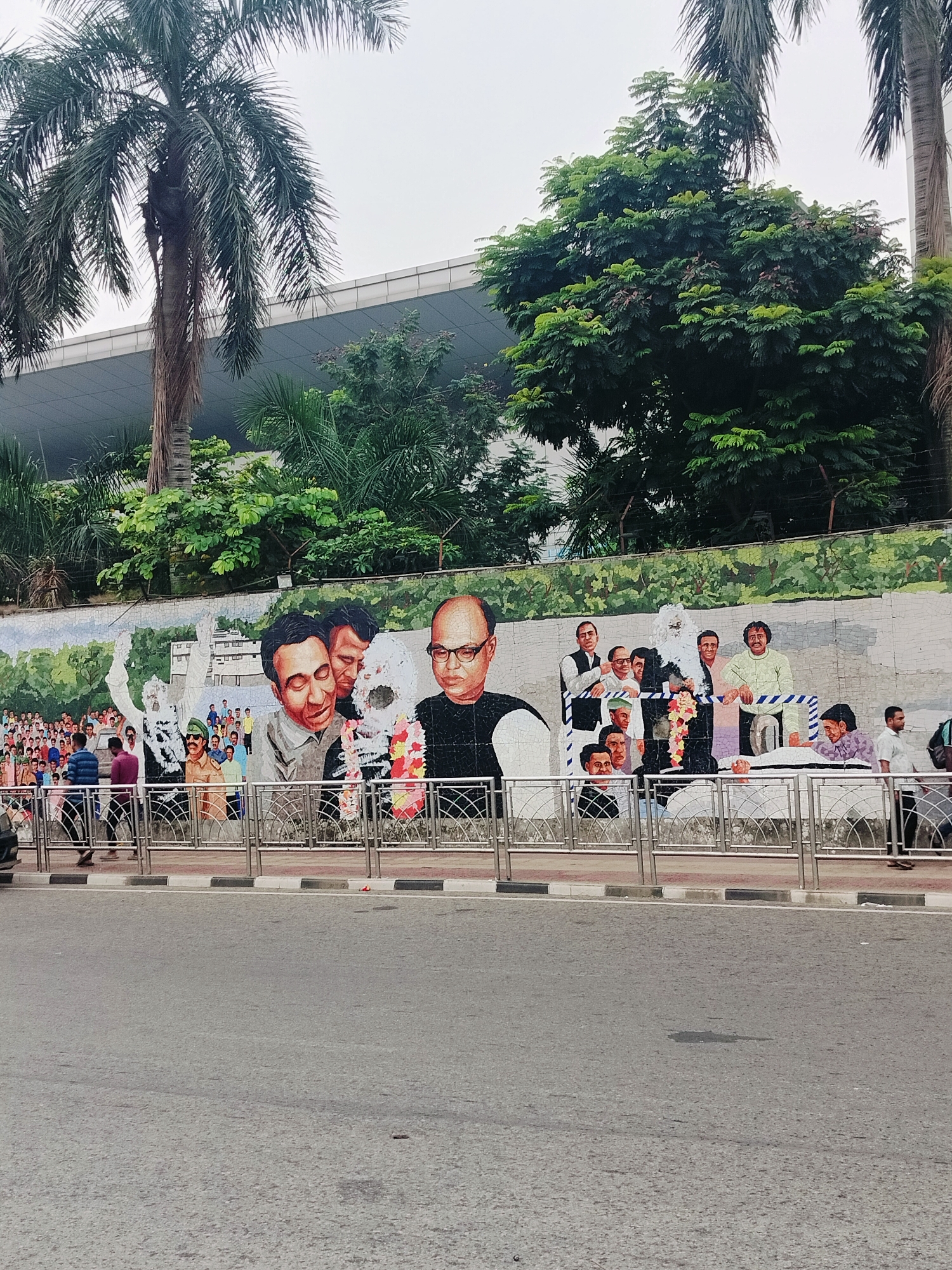 The image shows a large mosaic mural on a wall in Dhaka, Bangladesh, depicting a crowd of people and several prominent individuals. The mural is in a style that appears to commemorate historical figures or events, with large, stylized portraits of men in the foreground. Some of the figures are adorned with garlands, suggesting a celebratory or respectful occasion. Behind the main figures, a diverse crowd of smaller figures is depicted, rendered in a more abstract, mosaic style. The background of the mural features a green, hilly landscape with some buildings. In front of the mural, a silver metal railing separates the artwork from a paved walkway where several people are walking. Beyond the walkway, a road is visible with tire marks and some debris. Tall palm trees and lush green foliage are present in the upper portion of the image, with a modern building visible behind the trees. The sky is overcast.