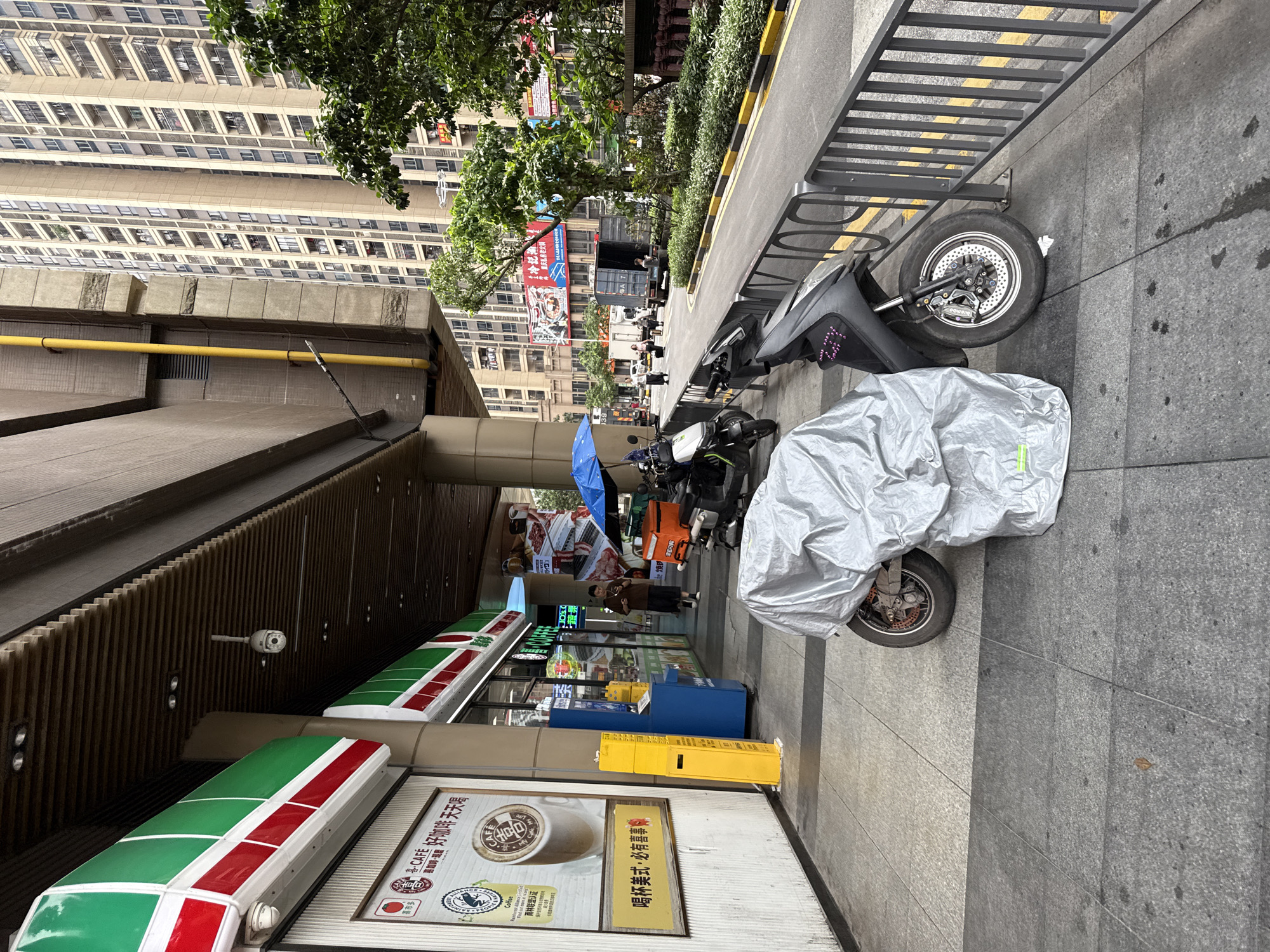 The image depicts an outdoor scene in Haizhu District, China, focusing on a sidewalk area in front of a convenience store. The store appears to be a 7-Eleven, identified by its distinctive green and red awning and signage. In the foreground, two parked electric scooters are visible, one of which is partially covered by a silver protective sheet. The scooter on the right is black and faces the street. A black metal fence separates the sidewalk from the road.
The sidewalk is paved with granite tiles. To the left of the scooters, a person in dark clothing walks away from the camera, towards the convenience store. The store features glass doors and windows, with displays of products visible inside. A yellow vertical vending machine or dispenser is positioned to the left of the store entrance. Above the store, a surveillance camera is mounted on the underside of a building overhang.
In the background, a modern, tall residential building with numerous balconies dominates the skyline. The street is asphalt and has yellow and black markings along the curb. Other elements in the background include trees, parked trucks, and more buildings. The overall impression is a typical urban street scene in China, with a focus on everyday commerce and transportation.