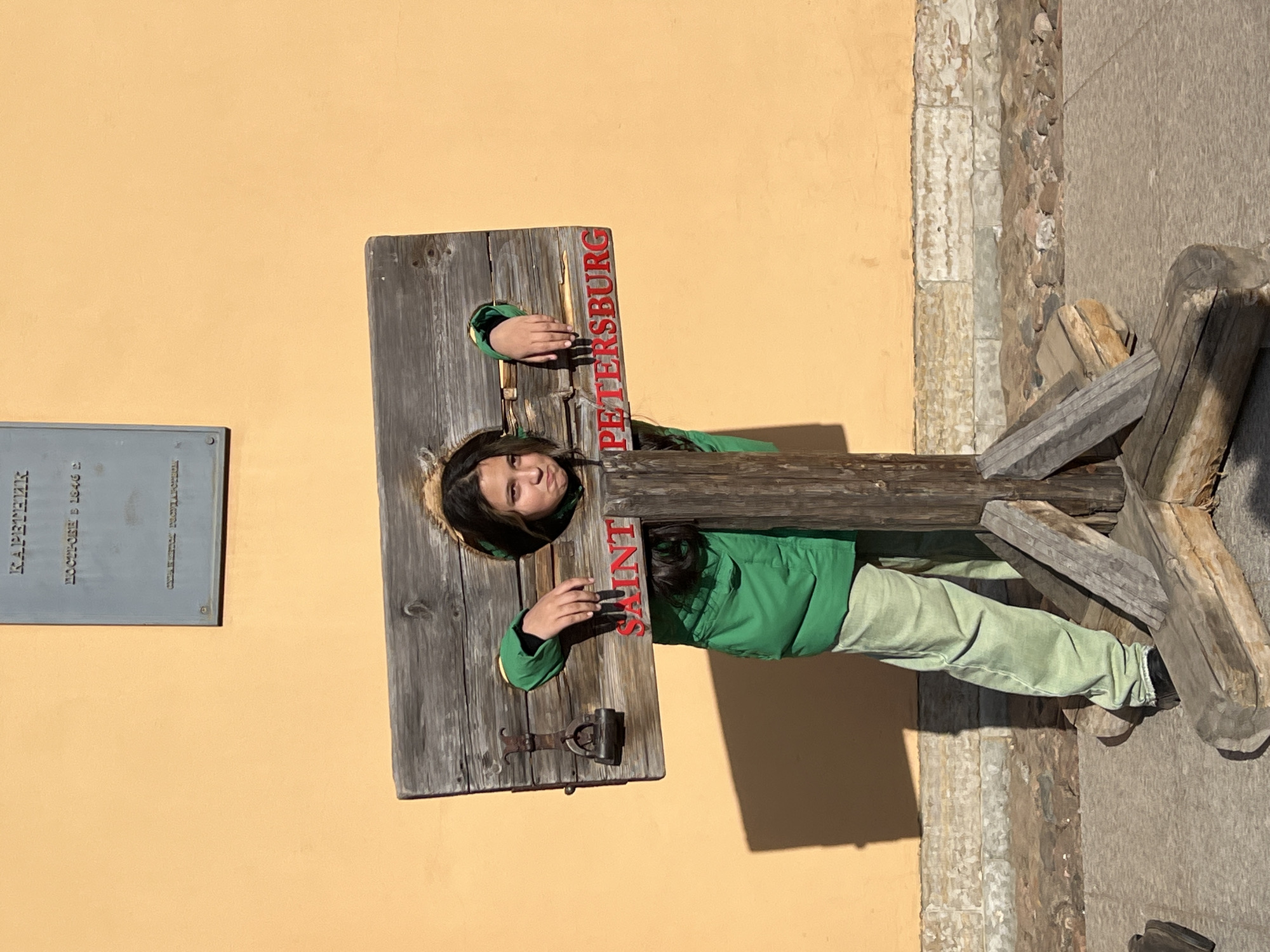A young woman in a green jacket and light green pants stands in a wooden pillory outdoors. The pillory has the words "SAINT PETERSBURG" written vertically in red letters. The pillory and a portion of a pale yellow building wall are on the left. A portion of a stone pathway is on the right. The sign on the building is written in Russian. The location is likely the Peter and Paul Fortress in Saint Petersburg, Russia.