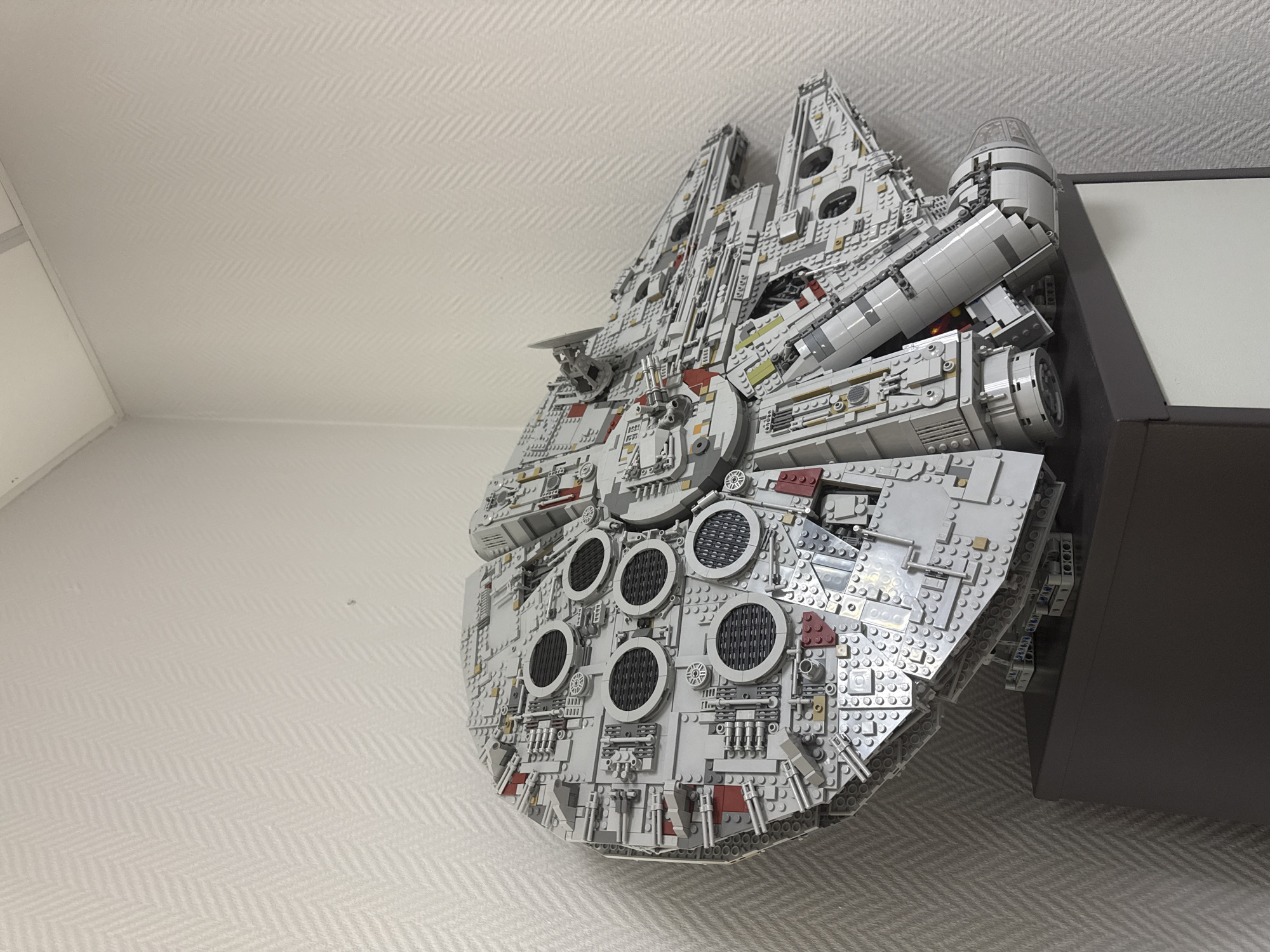 The image shows a large LEGO model of the Millennium Falcon mounted on a wall. The model is predominantly gray with intricate detailing. The wall has a light-colored, patterned wallpaper. The right side of the image includes a section of a dark-colored piece of furniture. There is no evidence to suggest this is in Krasnoyarsk, Russia, or any other specific location. There are no people visible in the image.