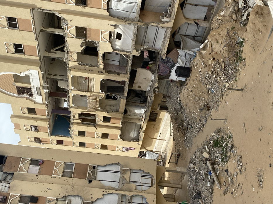 The image displays a severely damaged urban residential environment in Khan Yunis, Palestinian Territory, under an overcast sky. The foreground and midground are dominated by sandy, unpaved terrain extensively covered with large piles of concrete rubble, twisted metal rebar, and assorted debris. Two thin, dark posts protrude from the sand in the midground.
Two multi-story residential buildings, primarily light brown in color, show significant structural damage. The prominent building on the right exhibits catastrophic compromise, with multiple floors having entire sections of exterior walls and balconies completely missing, exposing the building's interior. Exposed areas reveal collapsed concrete slabs, damaged support beams, and various debris. Some interior spaces still contain visible household items, including clothes draped over railings. White and patterned tarpaulins are used as makeshift coverings over several exposed sections of the building's lower levels. The top floor of this building retains some original ornate arched architectural details.
To the left, another multi-story building, also light brown, presents numerous small, circular impact holes across its facade, consistent with bullet or shrapnel damage. Its lower levels are structurally compromised, with damaged walls and exposed interiors where white window blinds are visible. An arched entryway is partially visible at the base of this building.
A single individual, wearing a green garment, is partially visible walking on a sandy path in the lower-left corner of the image, moving from left to right. A black plastic barrel is located on the ground near the base of the severely damaged building on the right.