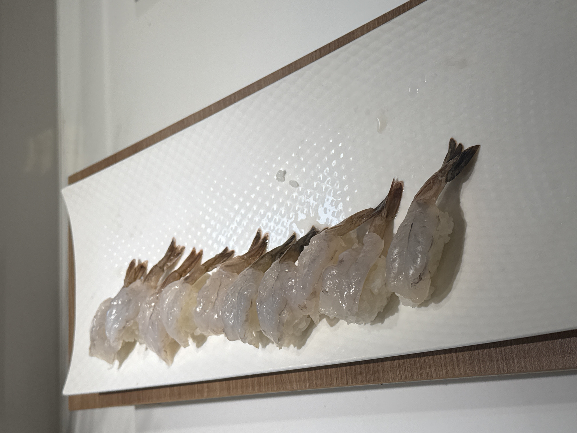 A close-up, high-angle, full-frame shot shows a white rectangular plate filled with raw shrimp sushi arranged in a line. The plate has a textured surface and is set on a light-colored countertop with a wooden border on the left side. Each piece of sushi consists of a small mound of white rice topped with a translucent, pale pink shrimp. The shrimp tails are visible, extending upwards and to the right. There are a few stray grains of rice and small droplets of liquid on the plate. The lighting is bright, casting soft shadows. The background is out of focus and appears to be a light gray wall and a white ceiling. The image does not contain any elements that suggest a specific location in Nha Trang Ward, Vietnam, or any actions or interactions.