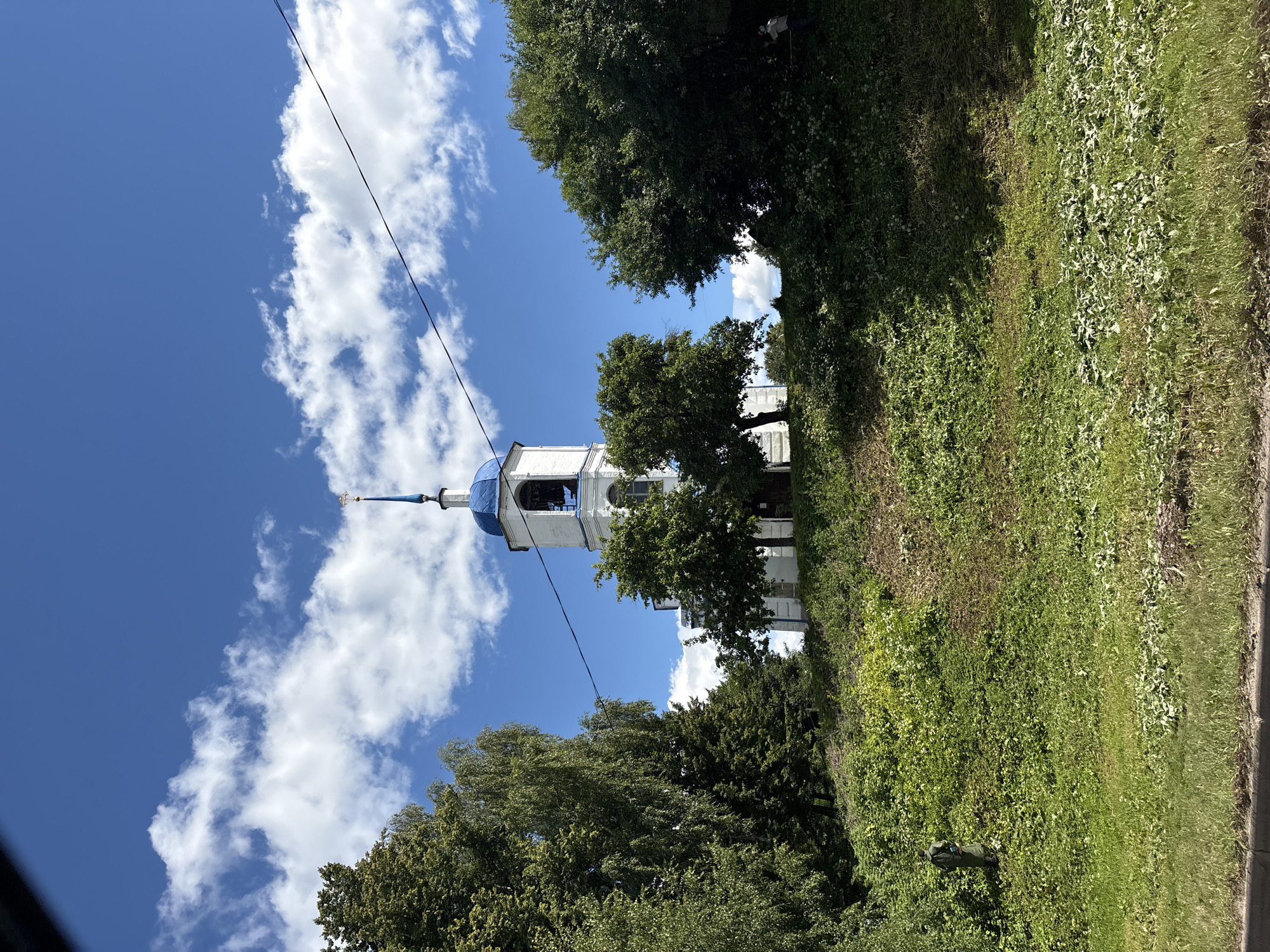 The image shows a white church with a blue domed belfry and a golden cross atop it, set against a bright blue sky with scattered white clouds. The church is situated on a grassy hill, surrounded by lush green trees. In the foreground, there is a slope covered in grass and various plants. A black electrical wire stretches across the upper part of the image, passing in front of the church. To the far right, a person in a blue shirt is visible, seemingly trimming vegetation with a tool. The overall scene appears to be a tranquil, daytime view of a religious building in a natural setting. The church depicted is likely the Church of the Transfiguration (Preobrazhenskaya Church) in Pereslavl-Zalessky, Russia, given its architectural style and the presence of a blue dome and belfry, common features in Orthodox churches of that region.