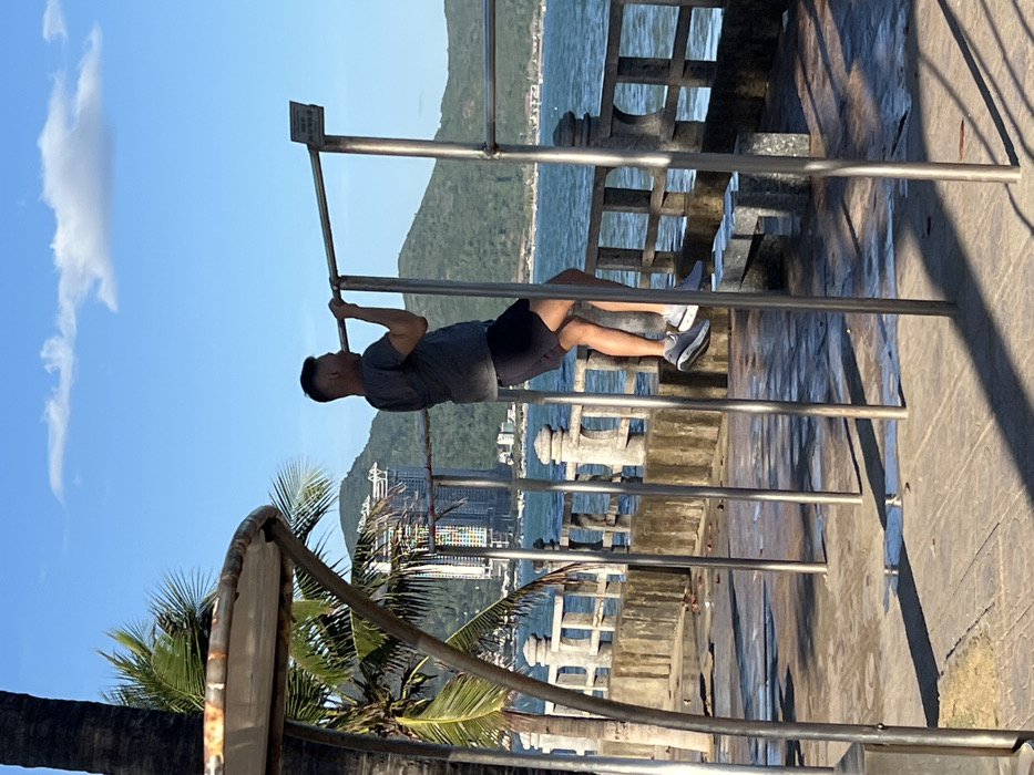 The image displays an adult male engaged in a pull-up on an outdoor stainless steel exercise bar station. The scene is set on a concrete promenade alongside a body of water in Nha Trang, Vietnam.
The subject, a male with short dark hair, is visible in profile, facing left, with his back partially to the viewer. He wears a grey short-sleeved t-shirt, dark athletic shorts, and grey and white athletic shoes. His arms are bent, and his body is elevated from the ground as he performs the exercise.
The exercise station consists of multiple vertical stainless steel posts supporting a horizontal pull-up bar. A small white rectangular sign with dark text is affixed to the upper right post. Long, sharp shadows from the equipment and an adjacent palm tree (whose trunk is visible on the far left with fronds extending into the upper frame) are cast across the concrete promenade, which appears to have wet patches.
Beyond the promenade, a decorative concrete balustrade separates the walkway from a calm, blue body of water, consistent with the South China Sea. In the mid-ground, across the water, forested green hills rise. Behind and partially obscured by these hills, multiple high-rise buildings are visible, including a distinctive modern structure with a colorful facade. The sky is clear blue with one elongated white cloud.
