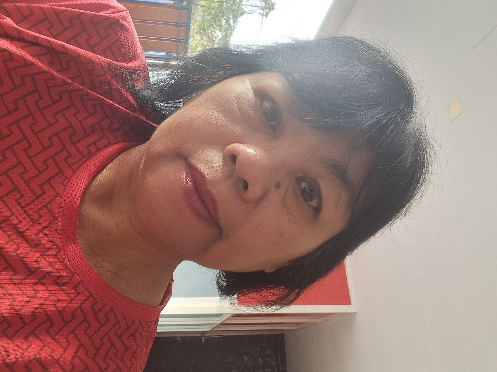 A close-up, slightly tilted selfie of a middle-aged East Asian woman is presented. She has dark, shoulder-length hair, dark eyes, and light skin. She is wearing a red t-shirt with a geometric pattern. The background includes a white ceiling with a square light fixture, and a red and white wall with vertical lines. To the left, a blurred outdoor scene with greenery and a fence is visible. The woman is positioned in the center of the frame, looking directly at the camera. The lighting suggests an indoor setting with natural light entering from the side. The image appears to be taken during the daytime.