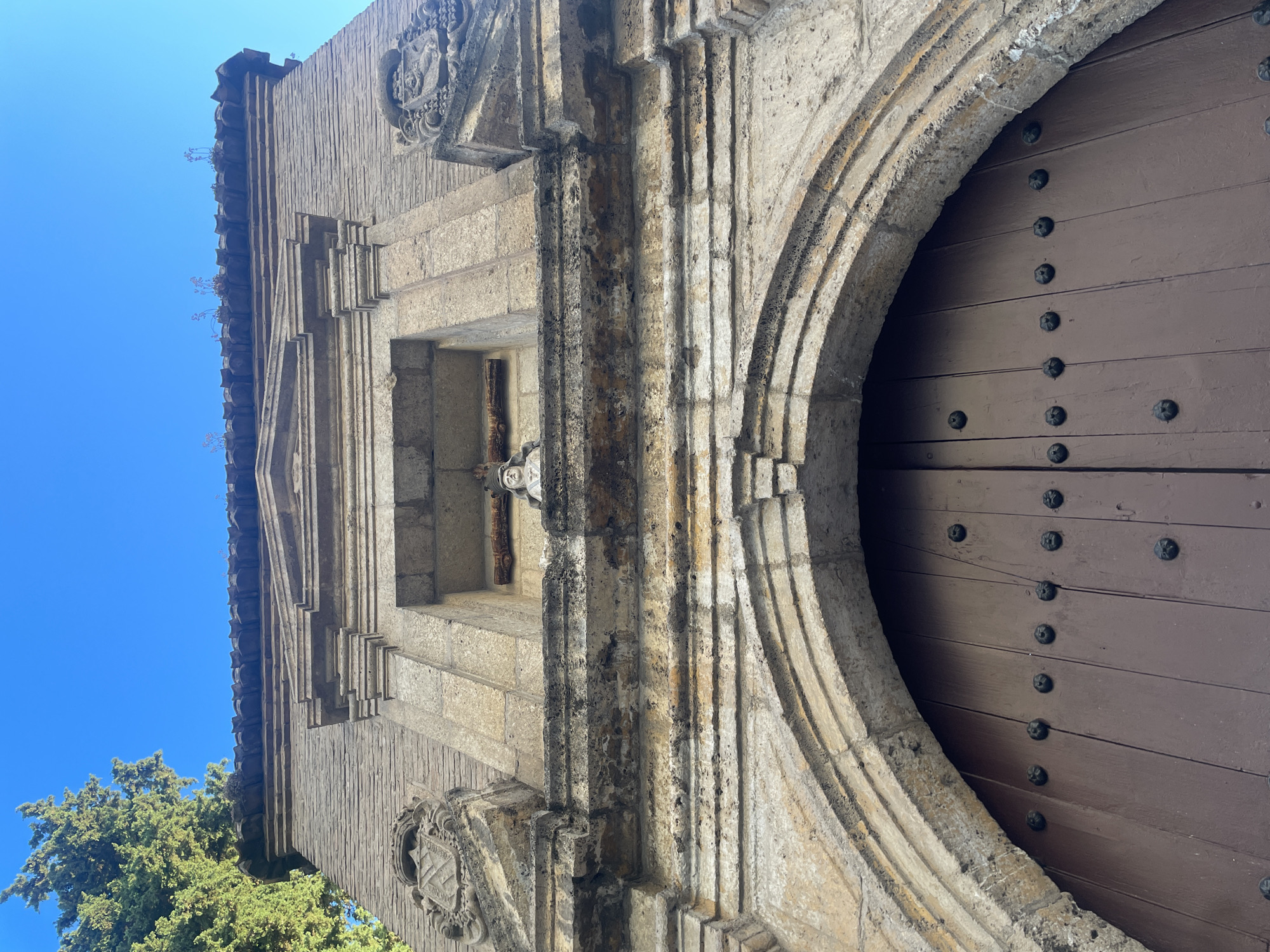 The image shows a close-up, low-angle view of the facade of a building, likely an entrance, in Granada, Spain. The upper portion of the image features a clear blue sky and the lush green foliage of a tall tree on the left. The building's facade is constructed of rough-hewn, light-colored stone and brick.
Dominating the lower part of the frame is a large, dark brown wooden door, divided into vertical panels and studded with numerous dark metal rivets. The door is set within a massive, arched stone entryway. The archway is intricately carved with concentric bands of stone, showing signs of age and wear.
Above the door and archway, the stone facade exhibits classical architectural details. There is a prominent horizontal stone ledge or frieze with carved decorative elements. Above this, a niche is visible, containing a small statue of a person wearing a helmet or crown and looking forward, with a wooden cross behind its head. Flanking the niche are two stone coat-of-arms with heraldic symbols. The roofline above the niche is adorned with terracotta tiles, some of which are cracked or missing, and small plants are growing from between them. The overall impression is one of historical architecture, suggesting a significant or old building.