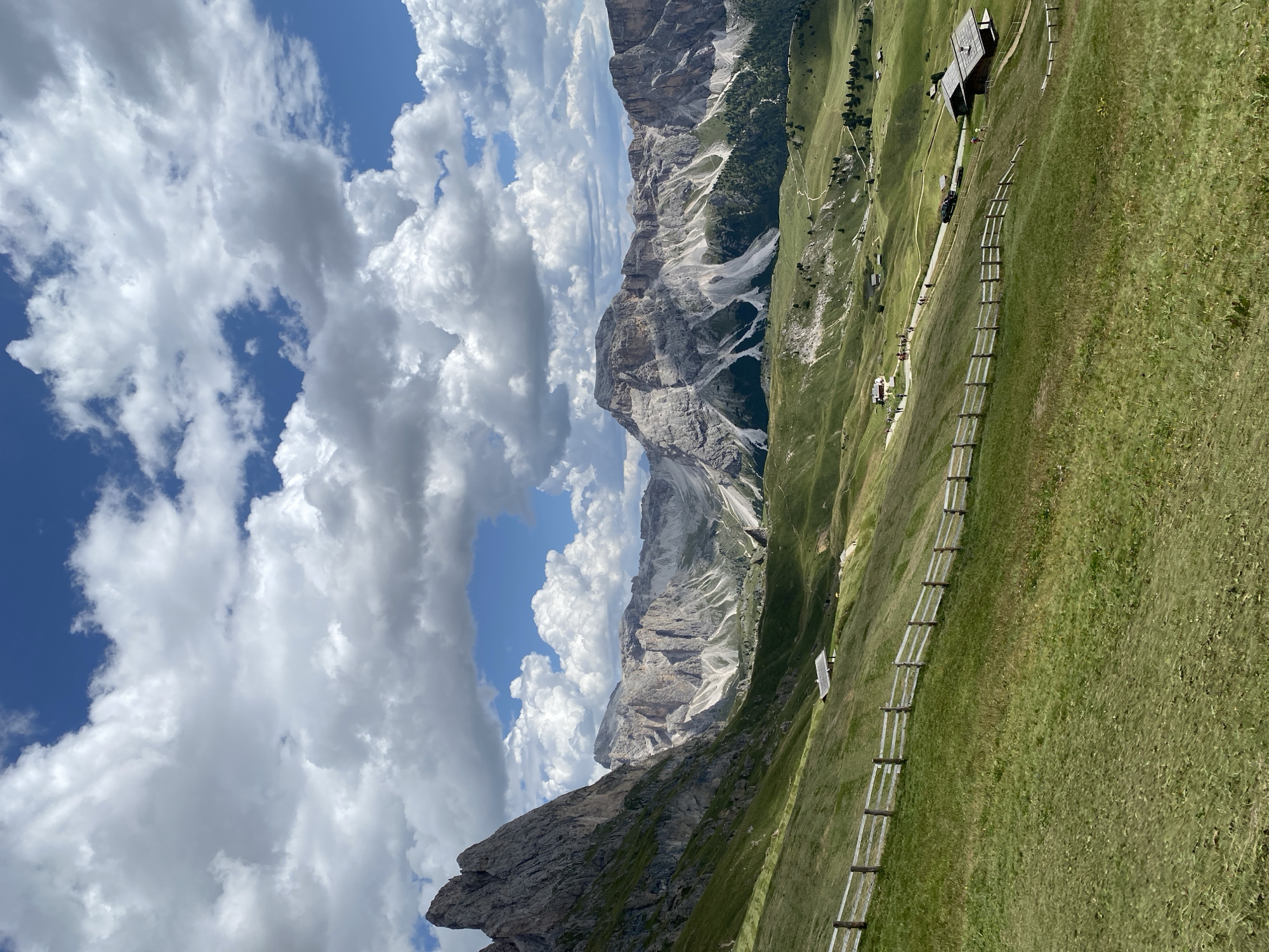 This is a landscape photograph of the Dolomites in Italy, specifically the Seceda ridgeline, under a partly cloudy blue sky. The foreground features a rolling green meadow with a rustic wooden fence running diagonally from the lower left to the middle of the frame. In the mid-ground, nestled into the hillside, are several small wooden buildings, one with a prominent roof structure. A paved road winds through the landscape, with several cars and a group of pedestrians visible. The background is dominated by the dramatic, jagged peaks of the Dolomites, with varying shades of grey rock and patches of green vegetation. The lighting suggests it is daytime, with the sun casting shadows and highlighting the contours of the mountains and the terrain.