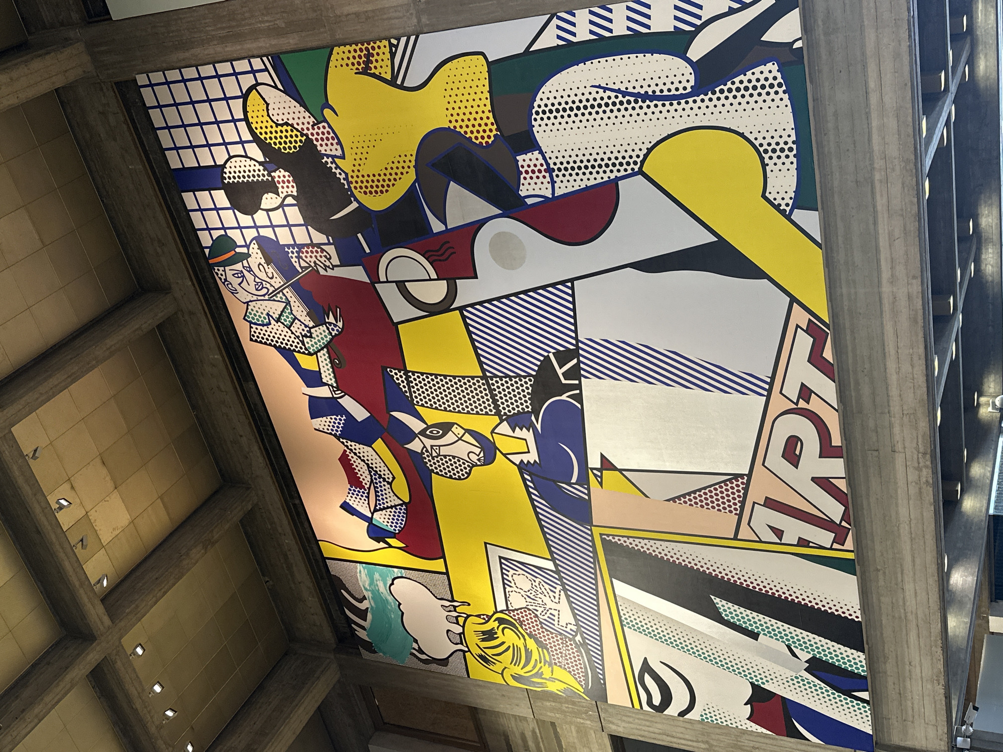 The image shows a large pop-art style mural on a ceiling within a building. The mural, reminiscent of Roy Lichtenstein's work, features various fragmented scenes and elements rendered in bold colors and Ben-Day dots. One segment depicts a man in a hat playing an instrument, while others include abstract shapes, faces, and the word "ART". The ceiling is part of a brutalist architectural structure with exposed concrete beams. Based on its known location, the image depicts the ceiling mural inside the Tel Aviv Museum of Art in Tel Aviv, Israel.