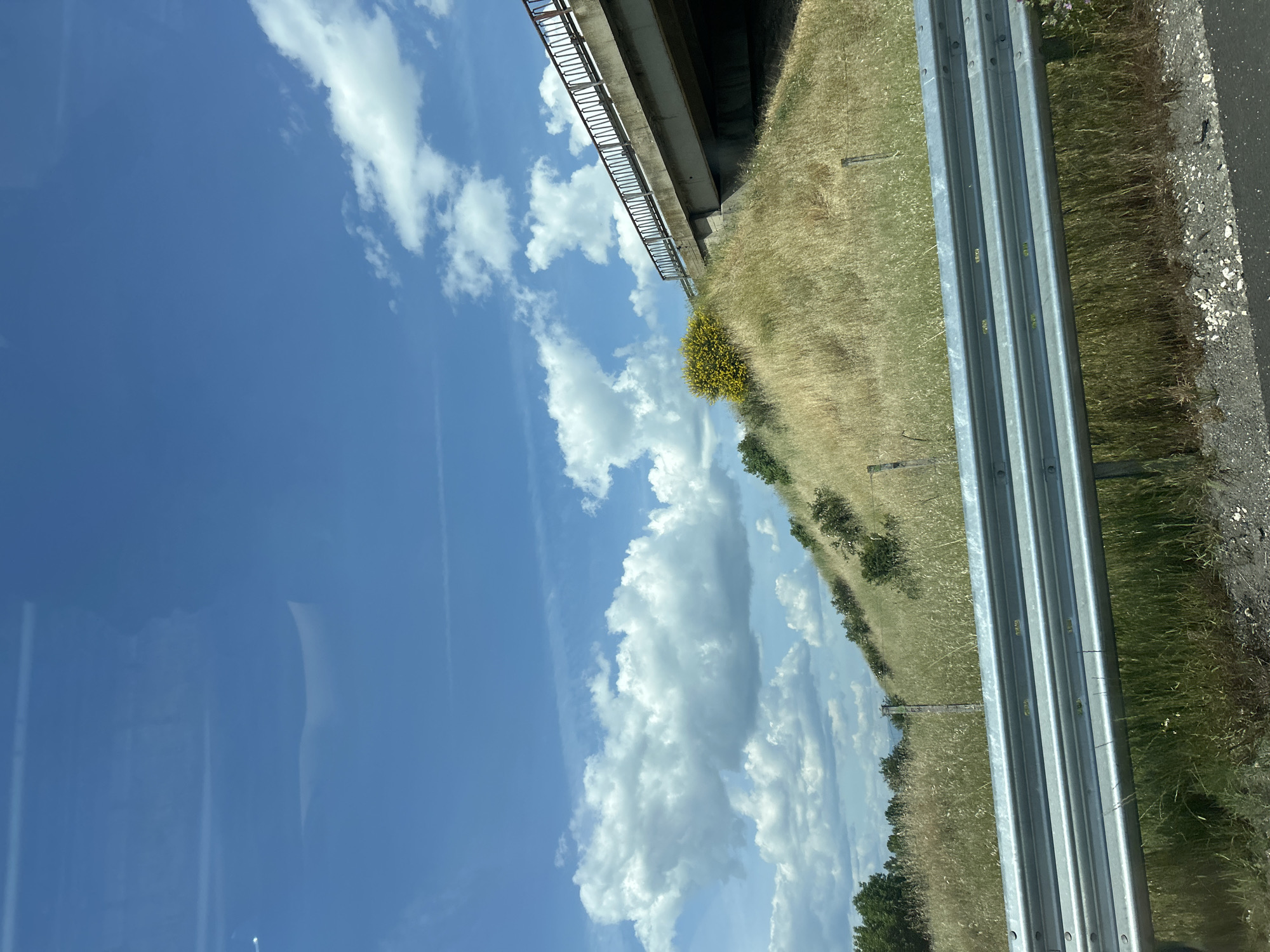 The image depicts a view from a moving vehicle, likely on a highway or roadway near Malo Konare, Bulgaria. The sky is predominantly blue with scattered cumulus clouds. A concrete overpass structure is visible above, casting a shadow on the grassy embankment. A metal guardrail runs along the right side of the road. The landscape features dry, grassy vegetation with sparse trees and shrubs. The road surface, composed of asphalt, is visible to the right of the guardrail.
