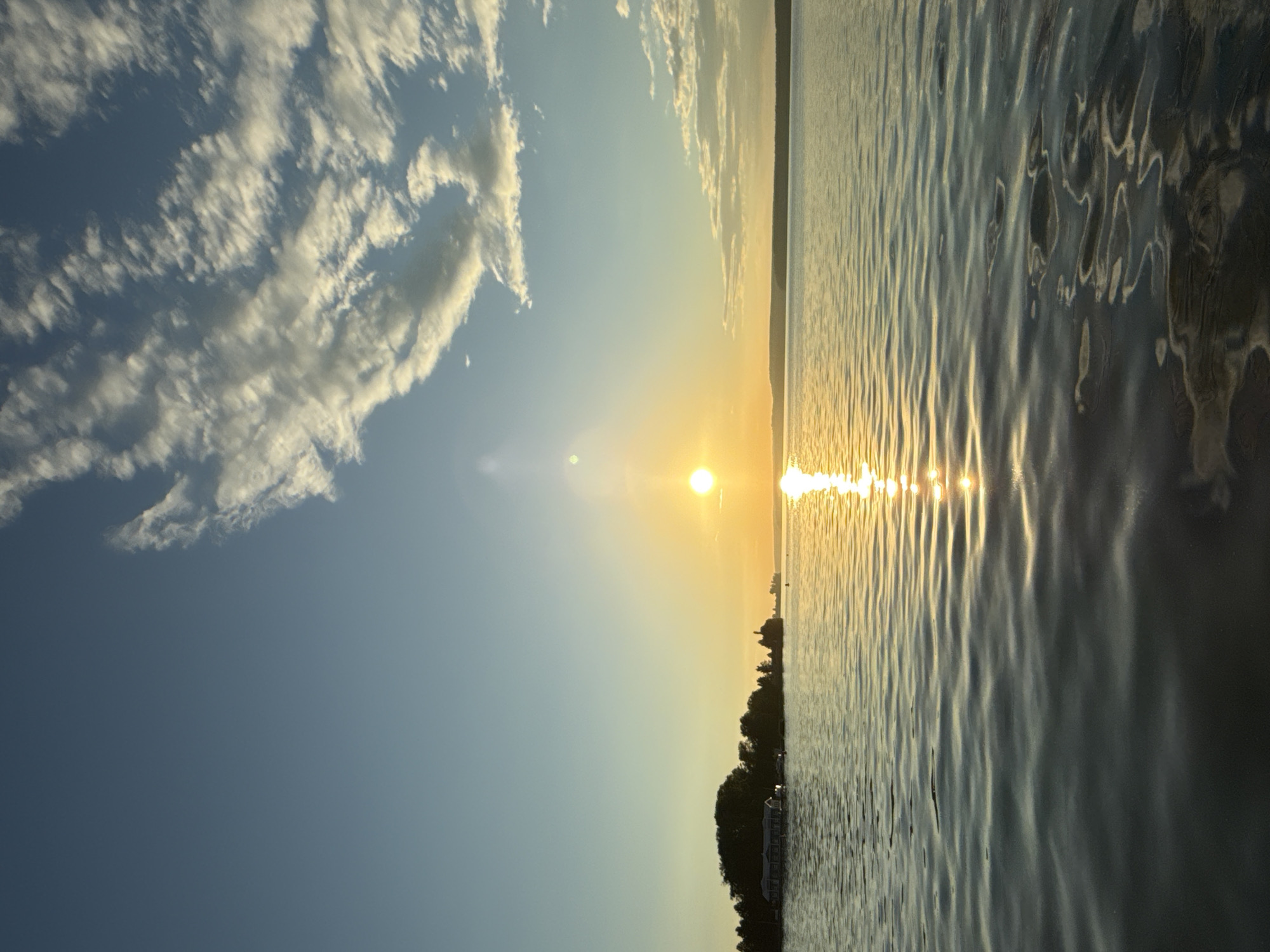The image depicts a serene sunset over a body of water, likely the Votkinsk Reservoir, a significant feature of Votkinsk, Russia. The sun, a bright golden orb, is positioned slightly left of center, casting a brilliant reflection on the rippling water's surface. The sky transitions from a pale blue at the top to a warmer yellow and orange hue near the horizon, where it meets a distant, tree-lined landmass. Wispy clouds, illuminated by the setting sun, are scattered across the upper portion of the sky. In the distance, on the left side of the frame, a small island or peninsula is visible, densely covered with trees and featuring a few structures, possibly buildings or boats. The foreground is dominated by the reflective water, showing gentle waves that create dynamic patterns of light and shadow. The overall scene conveys a sense of peace and natural beauty at the close of day. There are no people or animals visible in the frame.