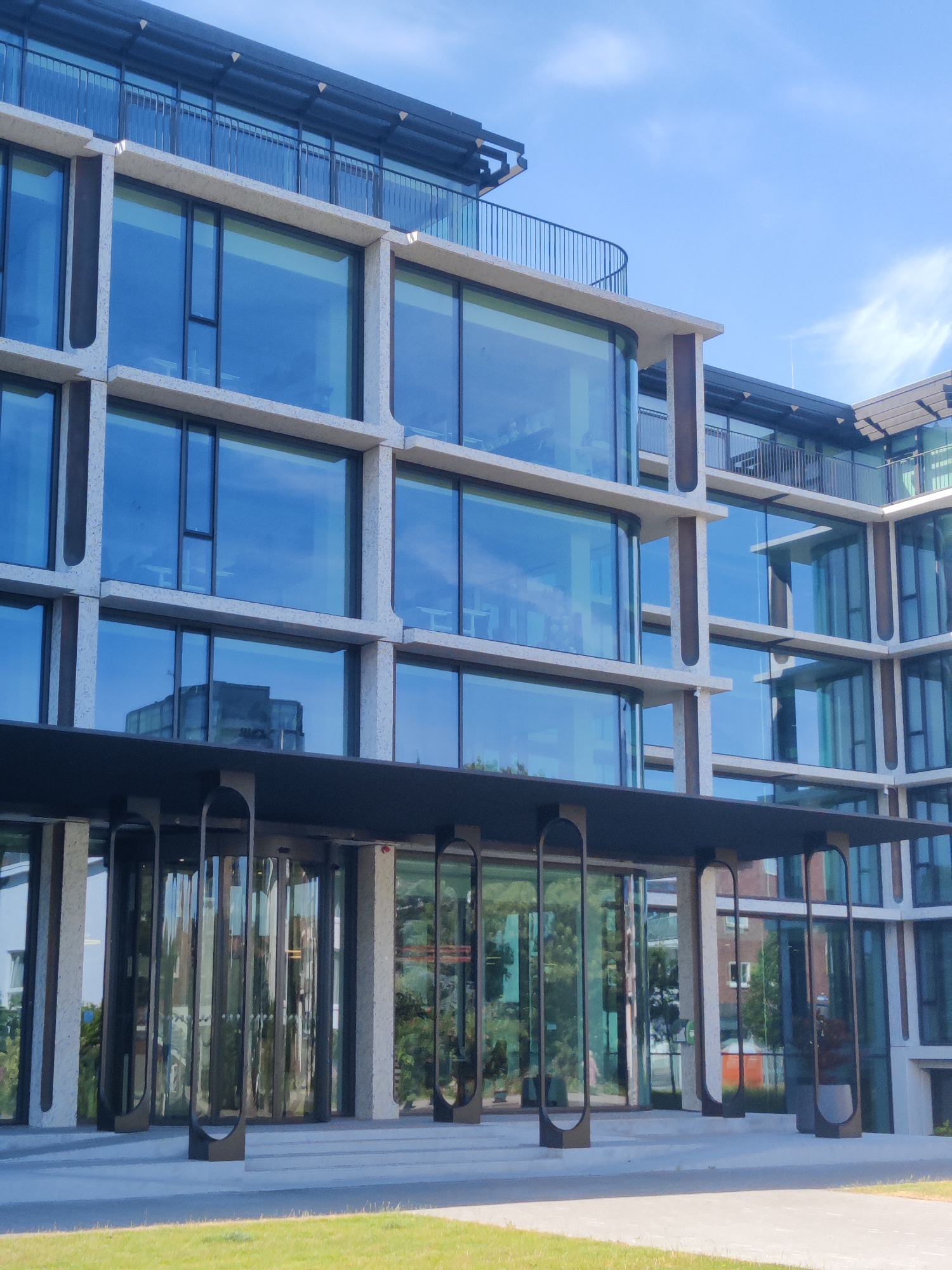The image shows the facade of a modern office building with a prominent use of glass and concrete. The building has multiple stories, each featuring large, reflective windows. The architectural style is characterized by horizontal bands of concrete framing the glass panes, creating a grid-like appearance. A curved balcony with a railing is visible on an upper level. The ground floor features a recessed entrance with a dark-colored canopy supported by vertical elements. The entrance area is equipped with revolving glass doors and vertical decorative frames. The building is situated in a well-maintained outdoor area with a green lawn in the foreground and a clear blue sky with sparse white clouds above. The reflection in the glass windows shows a neighboring building. The building is located in Amsterdam, Netherlands.