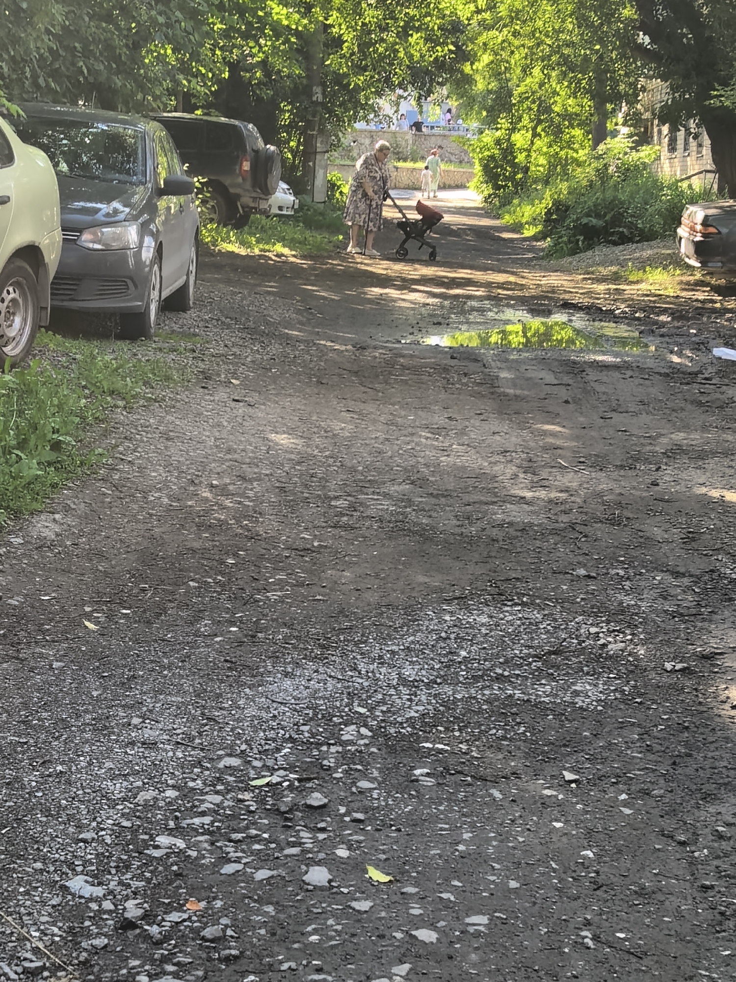 In an outdoor scene, a woman walks with a stroller on a dirt road. The road is uneven and shows signs of wear, with puddles of water reflecting the green foliage. Several parked cars are visible on the left side of the road, and a dark SUV is parked further down. Trees line the sides of the road, casting shadows and creating a dappled light effect. In the distance, a building with windows can be seen, and a few people are walking along a raised pathway. The environment appears to be a residential area or a park, with a sense of everyday life unfolding in a slightly unkempt setting.
