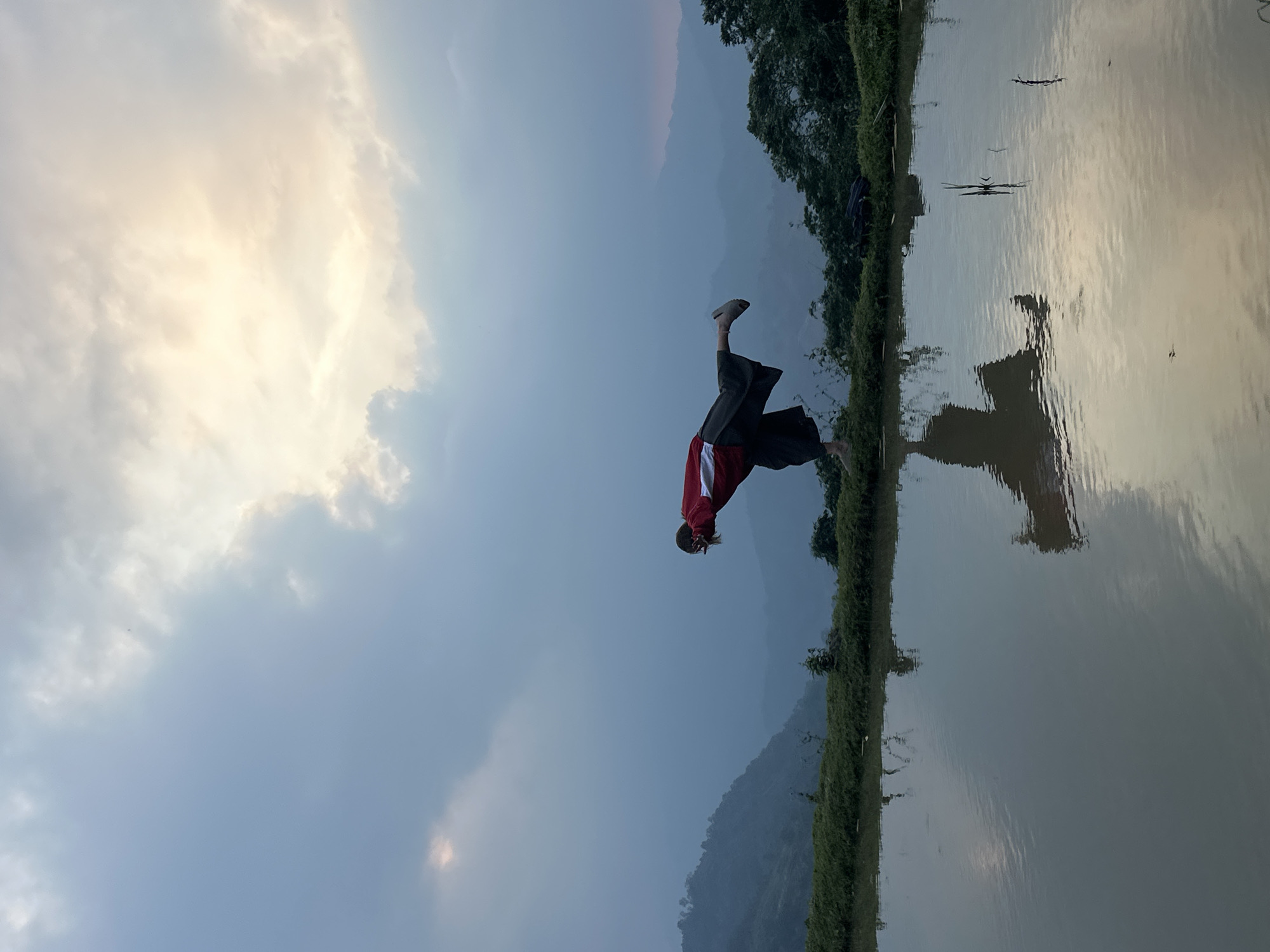 This image captures a person in an inverted acrobatic pose by a calm body of water in a natural, serene setting, identified as Nậm Dịch Commune, Vietnam.
The main subject is an individual, seen performing what appears to be a handstand or a similar inverted maneuver. They are wearing dark trousers, a long-sleeved top with red and white sections, and light-colored shoes. Their light-colored hair is pulled back. The person is positioned very close to the water's edge, and their complete reflection is visible in the still water.
The scene features a wide, placid body of water that acts as a mirror, reflecting the sky, the person, and the distant landscape. Along the left bank of the water, there is a narrow band of lush green vegetation, likely bushes or small trees. Beyond this greenery, the terrain rises into hazy, forested mountains or hills that recede into the distance. The sky above is largely cloudy, with soft, bright patches indicating the presence of the sun, casting a gentle, warm light over the scene.
The lighting suggests either an early morning or late afternoon, possibly sunset, creating a soft and atmospheric glow. The weather appears calm and partly cloudy. There is no visible text in the image.