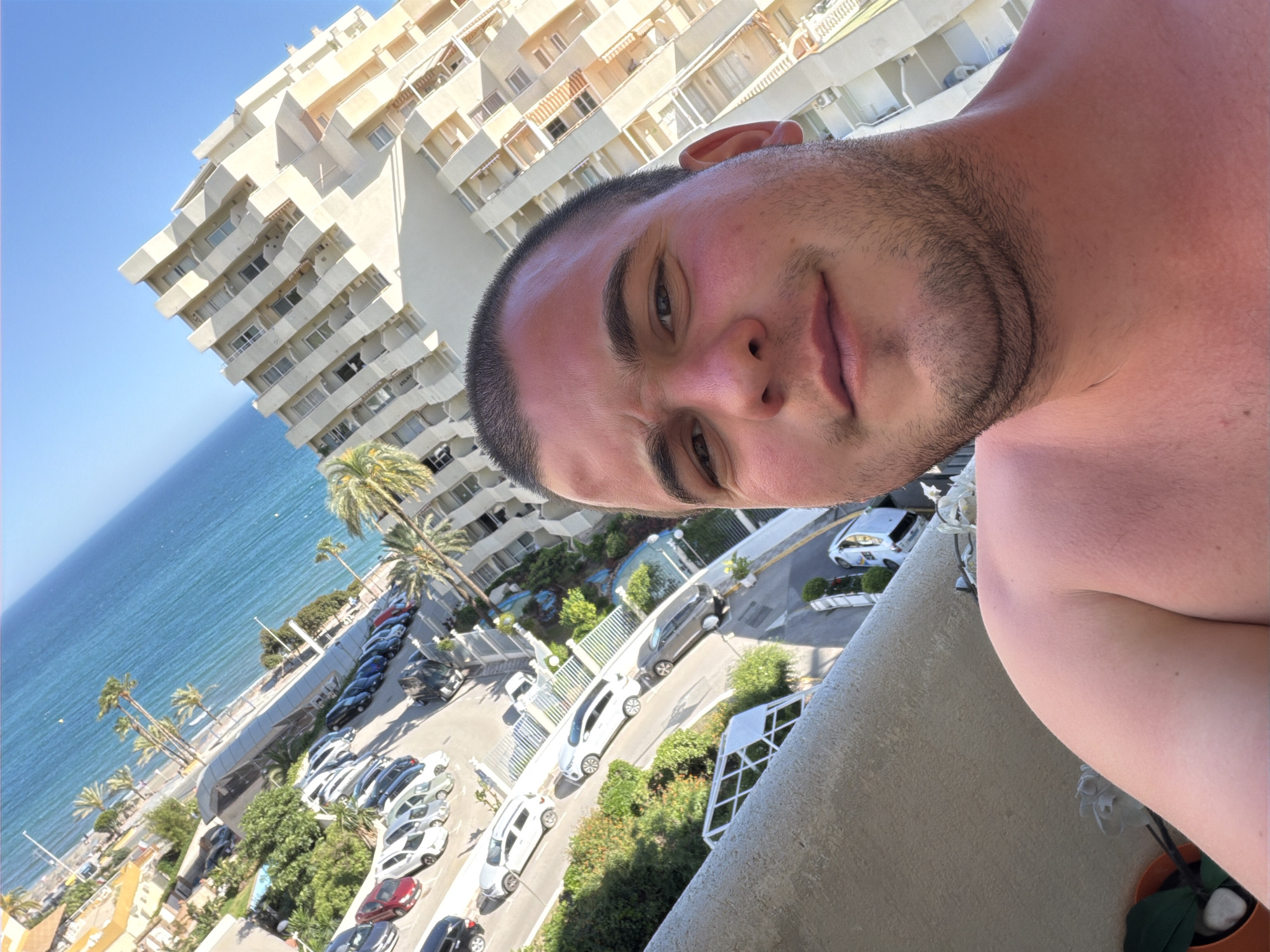 A selfie-style photograph is taken at an angle, featuring a man's face and upper body, with an urban coastal setting in the background. The man has short, dark hair and a stubbled beard. He is fair-skinned, and his chest and shoulders are visible.
The background includes the ocean, a beach with palm trees, a parking lot with cars, a road, and multi-story buildings. The architecture includes apartment buildings with rows of balconies.
Based on the architecture, urban layout, and proximity to the sea, the location is identified as Arroyo de la Miel-Benalmádena Costa, Spain.